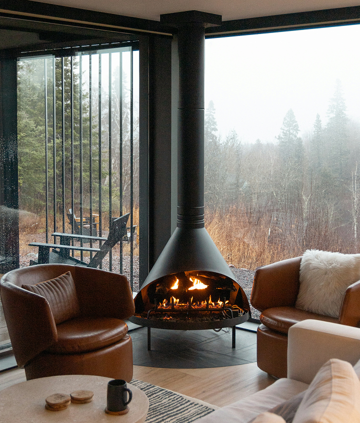 Living room with modern fireplace, brown leather chairs, white sofa with fur pillow, round coffee table with a mug, and large window showing trees and outdoor seating.