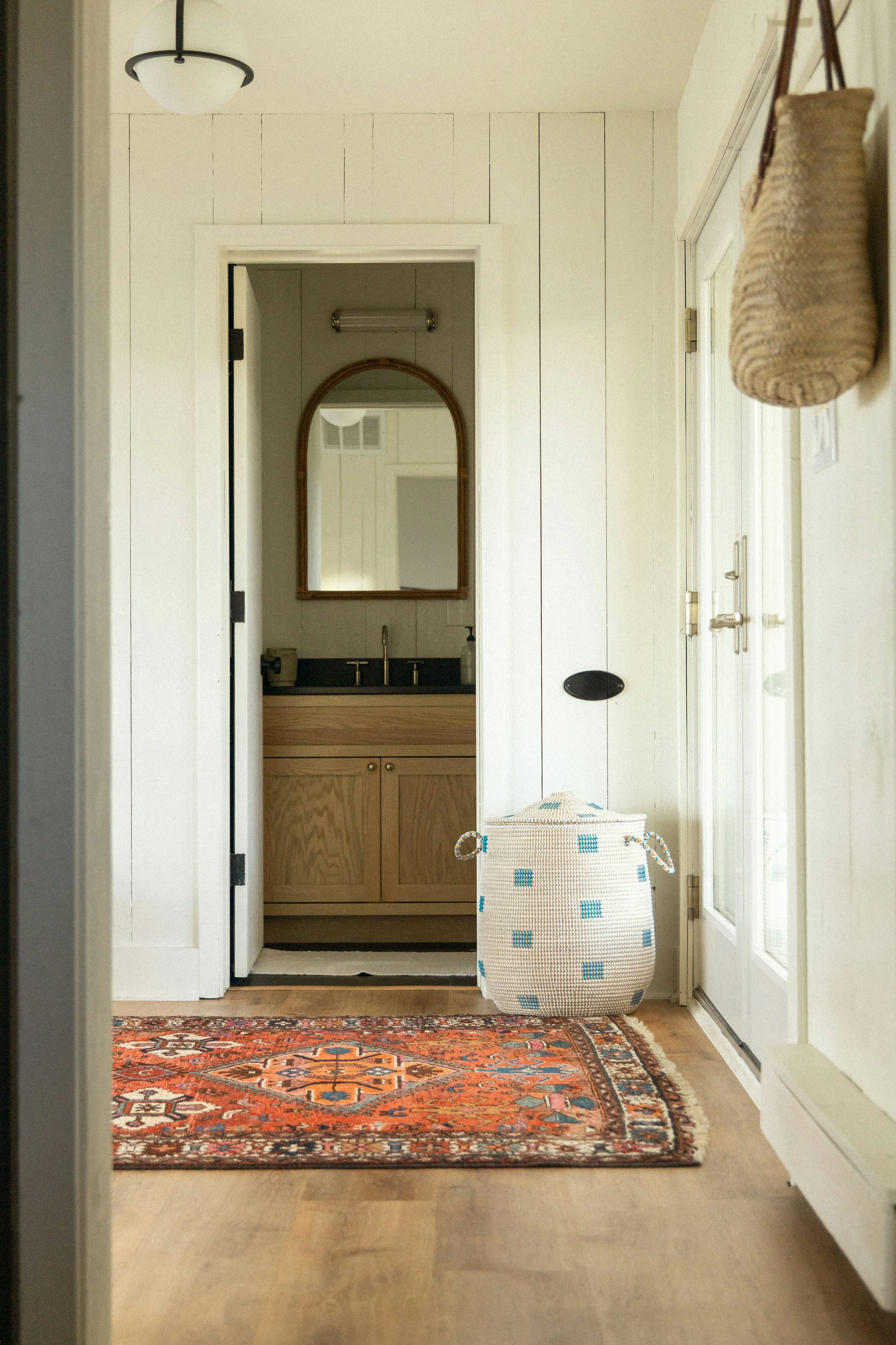 Interior view of a porch or entryway with a colorful oriental rug, a large woven basket with blue squares, and a sliding glass door leading outside. There is a small black oval sign or plaque on the wall, and a door with a handle on the right side.
