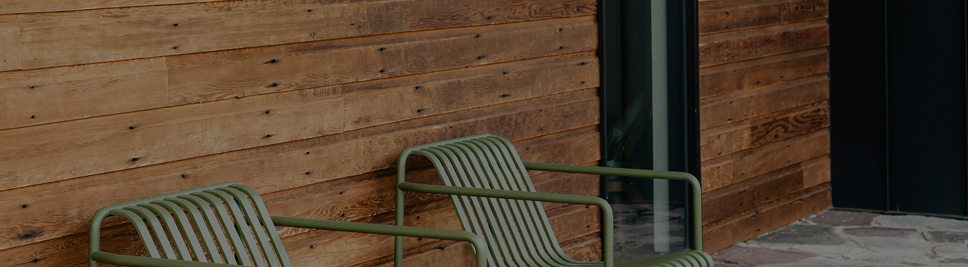 Two green metal chairs with slatted seats and backs placed against a wooden wall outside a building.