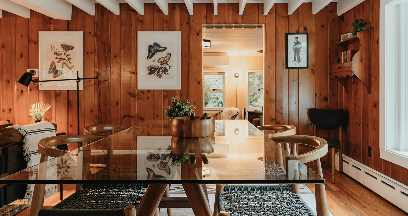 A cozy dining room with wooden paneled walls, a glass-top table, and six wooden chairs. There are framed butterfly and bird art prints on the wall and a window letting in natural light.