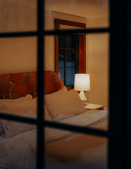 A cozy bedroom seen through window blinds, featuring a bed with pillows, a swan-patterned headboard, a window with wooden trim, and a bedside lamp emitting warm light.