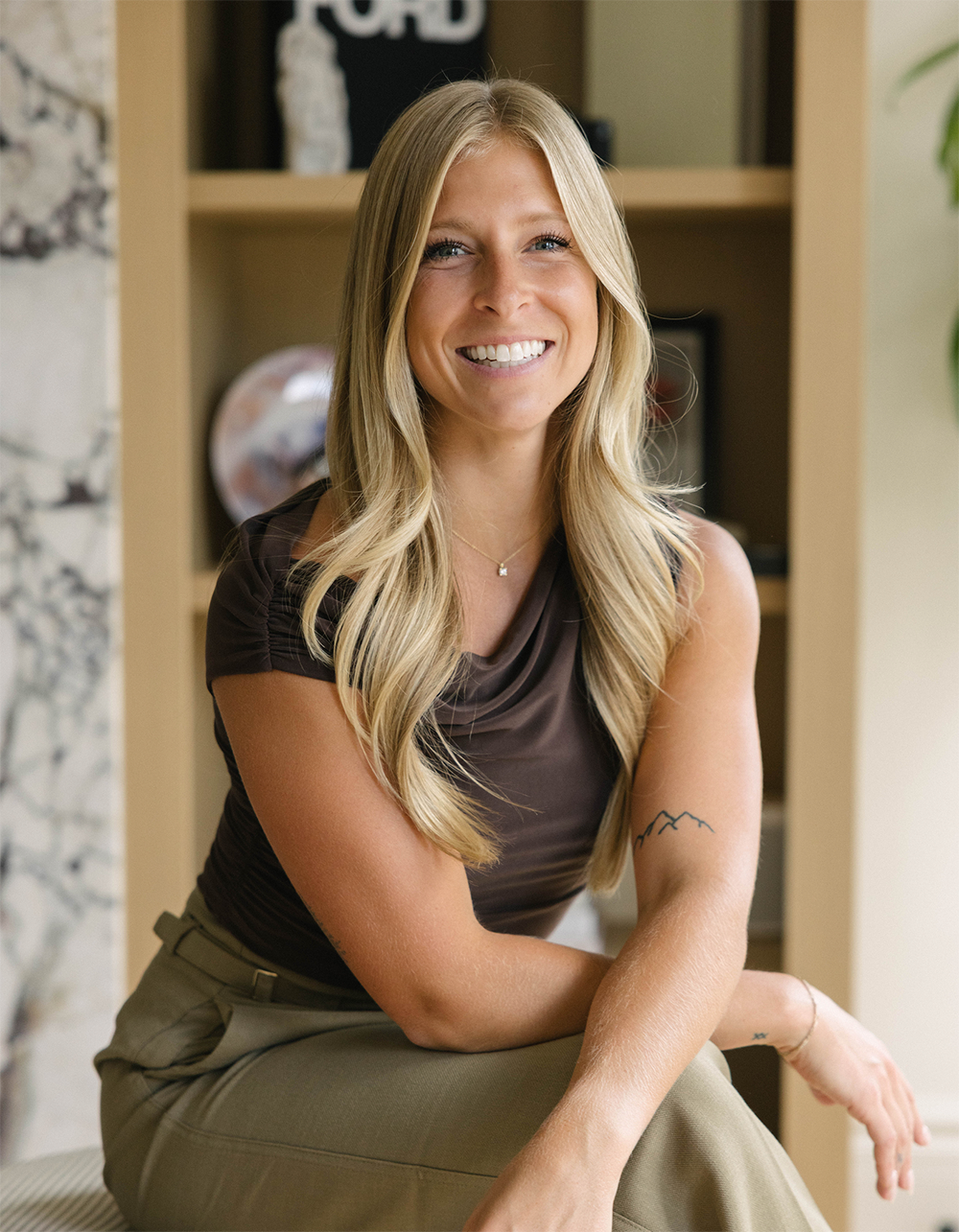 A smiling woman with long blonde hair, wearing a dark brown top and khaki pants, sitting indoors in front of a bookshelf.