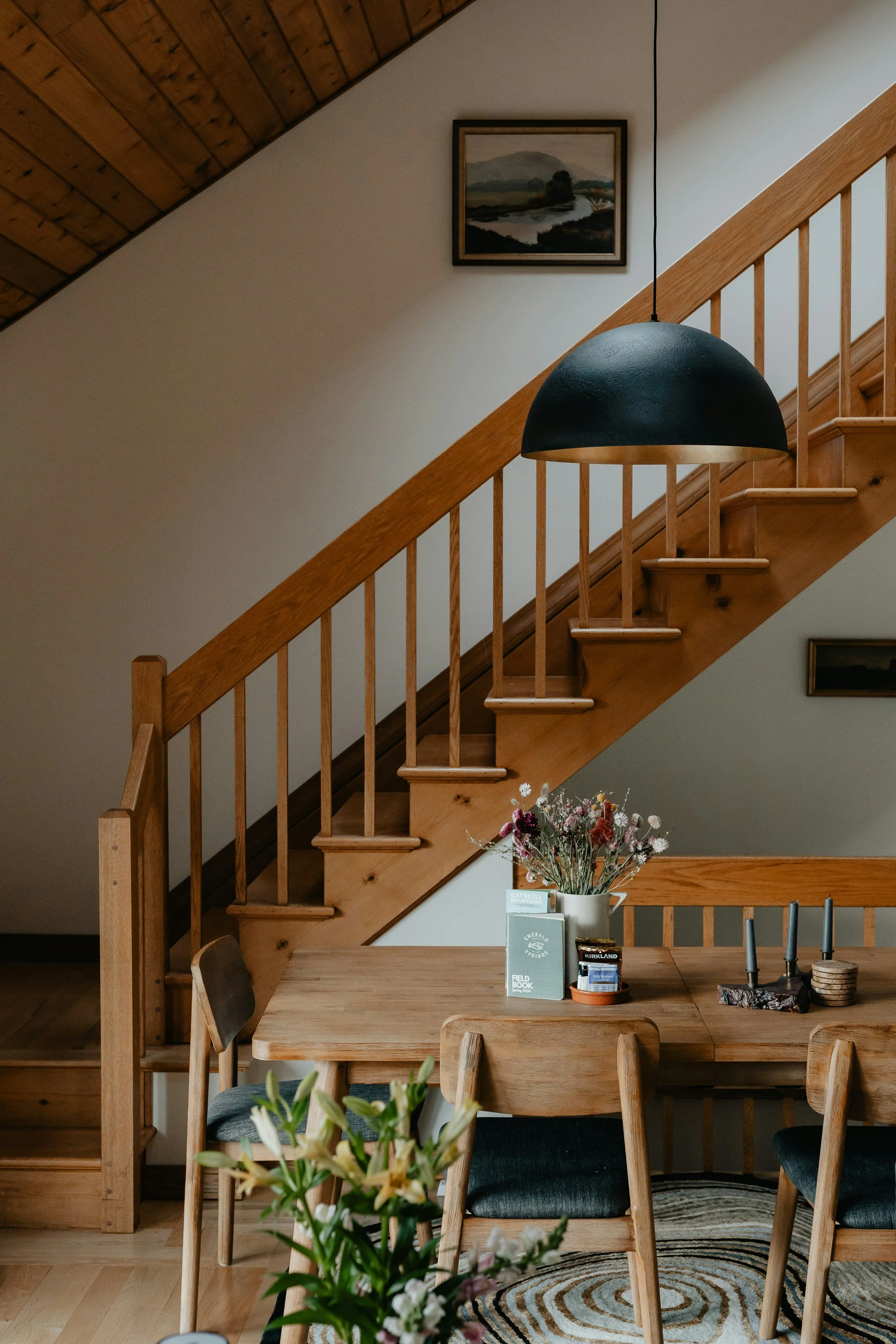 Interior of a home featuring a wooden staircase with a painting of a river landscape hanging on the wall above it. A black pendant light hangs over a dining table that has a flower arrangement, books, and candleholders on it. Part of a wooden dining chair and a rug are visible in the foreground.