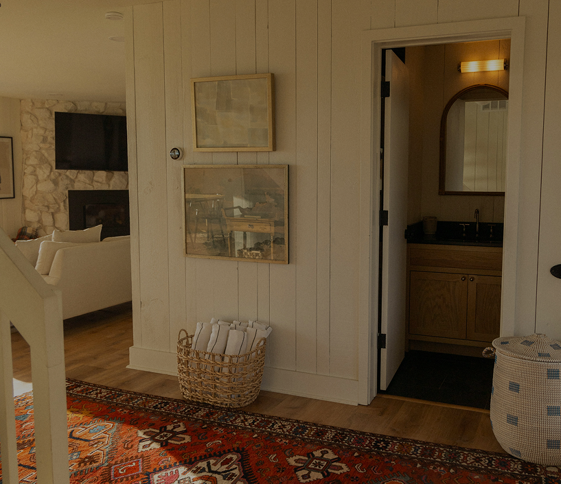 Interior view of a cozy living space with a white wooden wall, two framed pictures, a basket with rolled towels, a doorway leading to a bathroom with a mirror and sink, a patterned area rug, a white couch, and a stone fireplace with a mounted TV.
