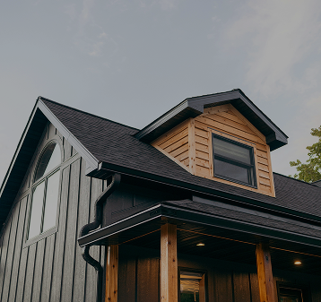 Close-up of a modern house's roof and upper facade, featuring a gable roof, black siding, wood accents, and a large window.