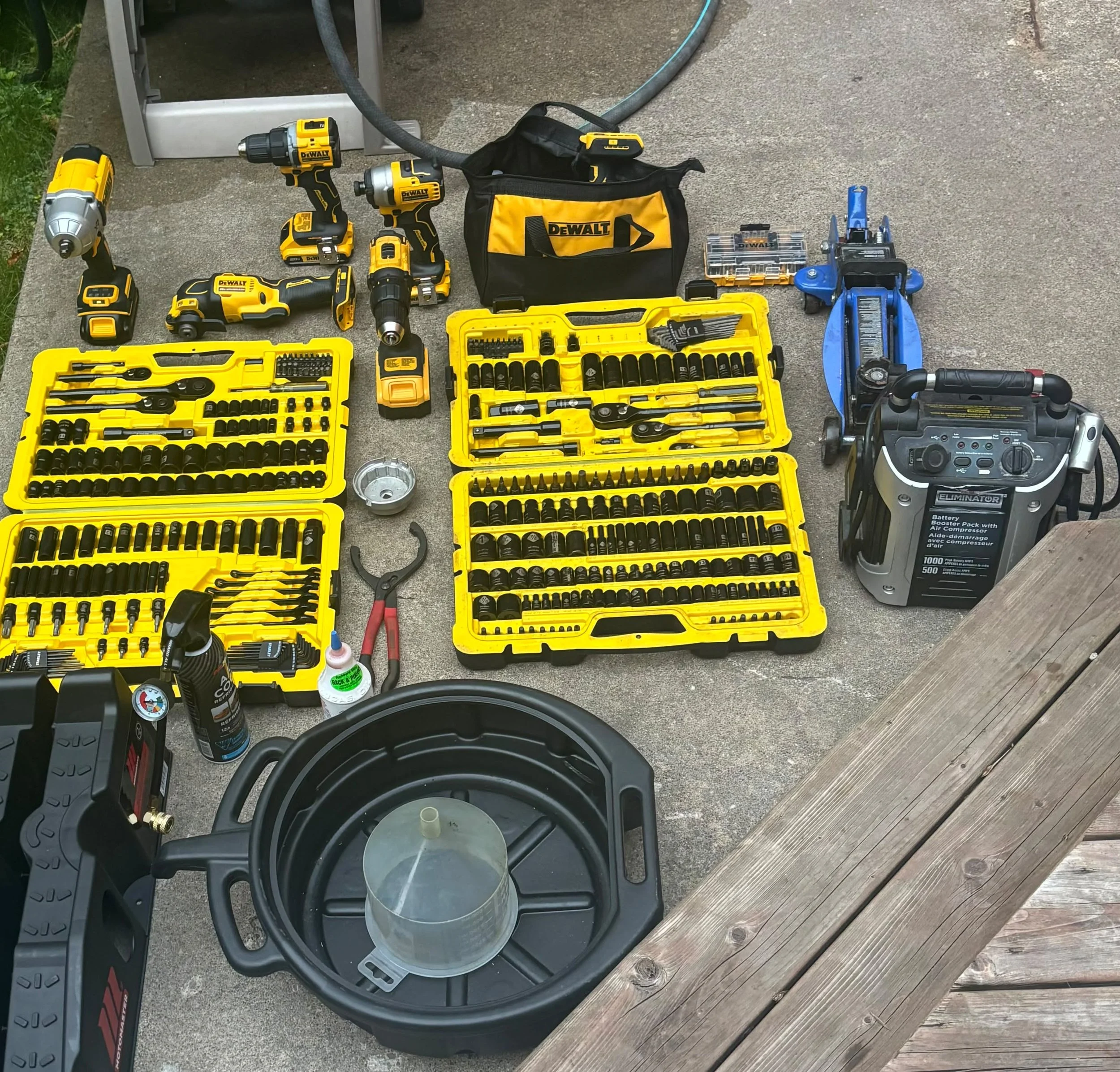 Tools and equipment laid out on concrete and wooden deck, including cordless drills, a toolbox, a handheld power saw, a cordless impact wrench, a cordless screwdriver, a bottle of adjusting oil, a saw blade, a battery charger, and a large toolset in yellow cases, along with a black toolbox bag with a yellow Dewalt logo.
