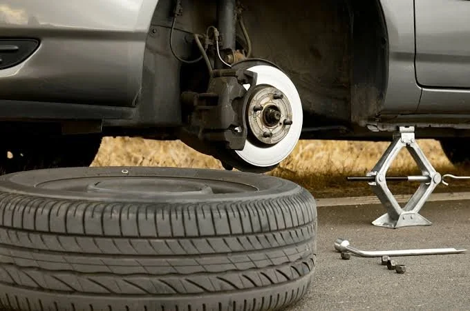 A car with a wheel removed, supported by a jack stand; a spare tire and tire removal tools are on the ground nearby.