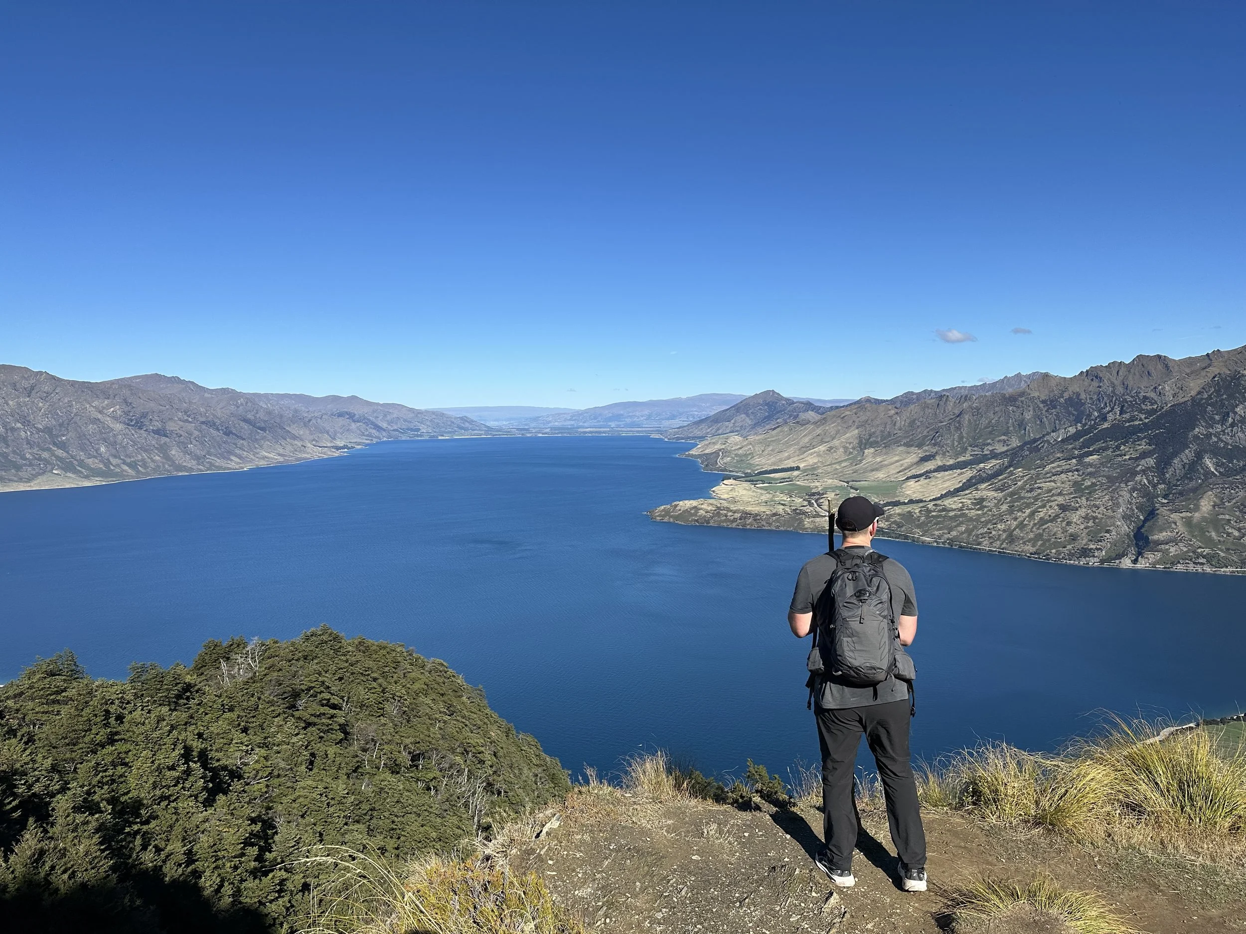 A person wearing a black cap, gray shirt, and black pants with a backpack stands on a dirt path overlooking a large body of water with mountains on the far side under a bright blue sky.
