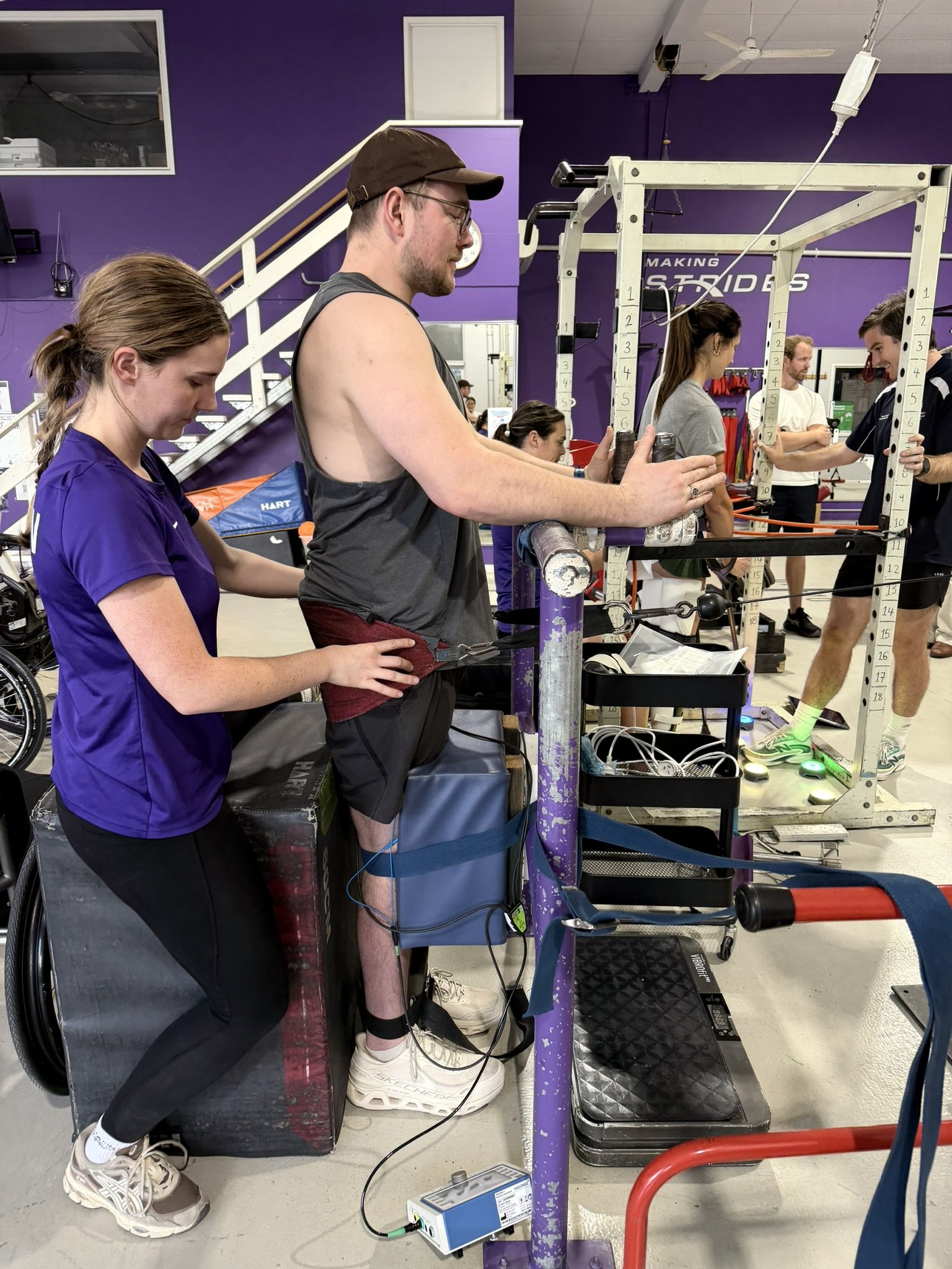 Person on a treadmill in a gym getting fitted with motion sensors by a staff member, with other people in the background.