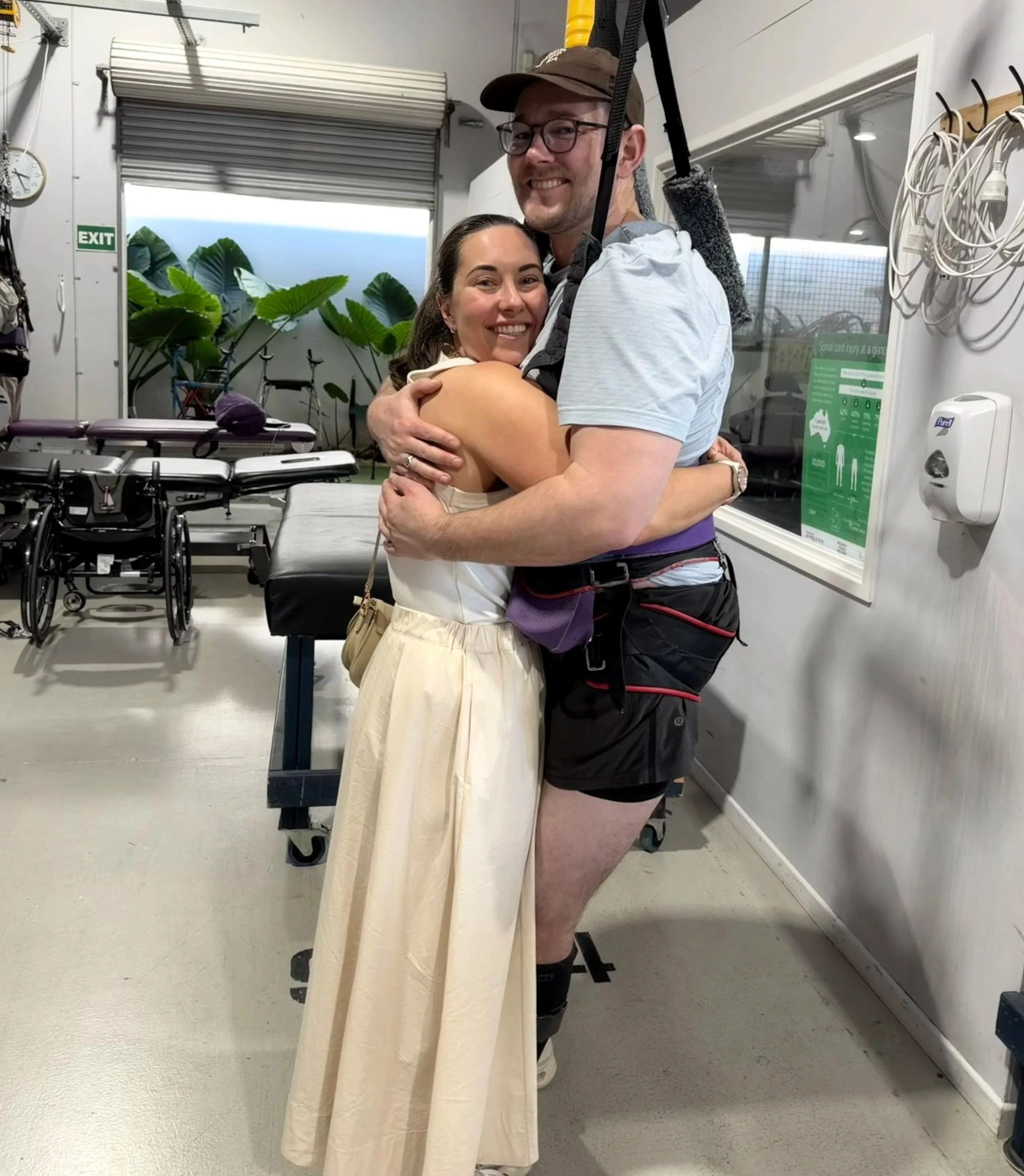 A man and woman embrace in a hospital or medical facility, with wheelchairs and medical equipment visible in the background. The woman is wearing a light dress, and the man is dressed casually with a harness or climbing gear over his shoulder, smiling at the camera.