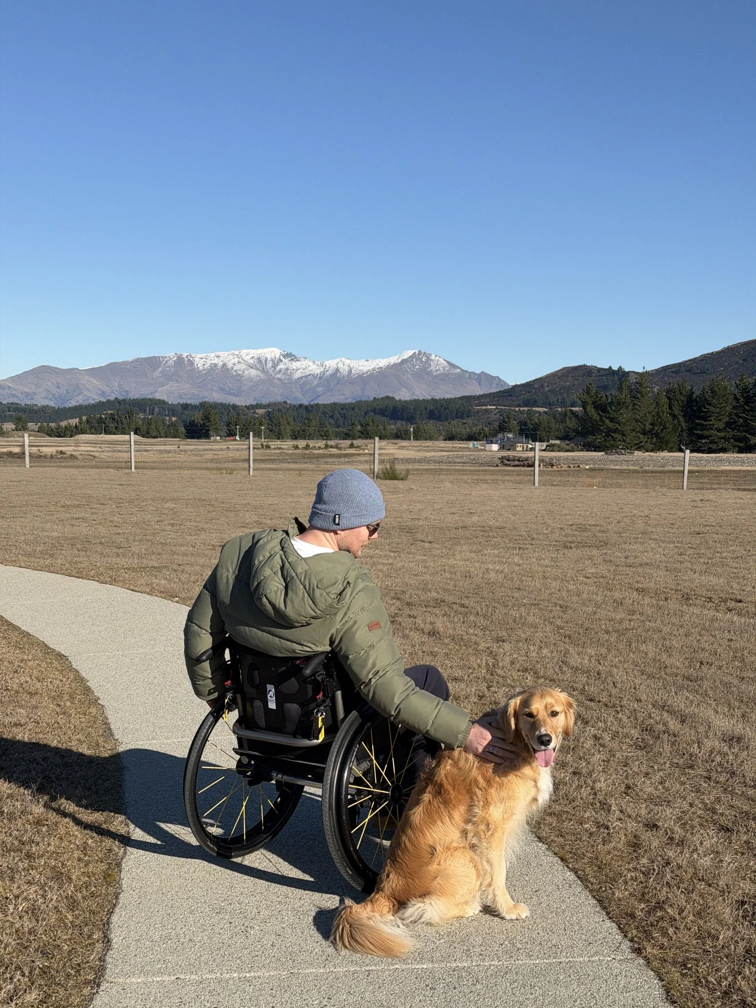 A man in a wheelchair petting a golden retriever dog on a paved sidewalk near an open field with mountains in the background under a clear blue sky.