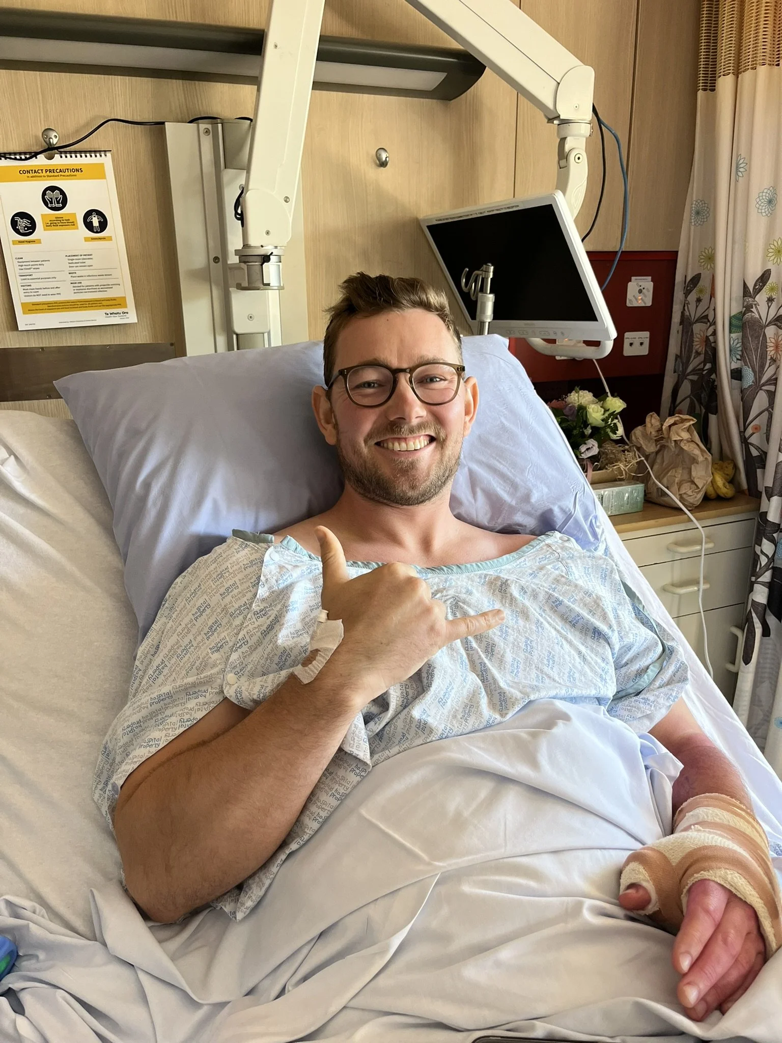 A young man with glasses lying in a hospital bed, smiling and giving a thumbs-up, with medical equipment and flowers in the background.