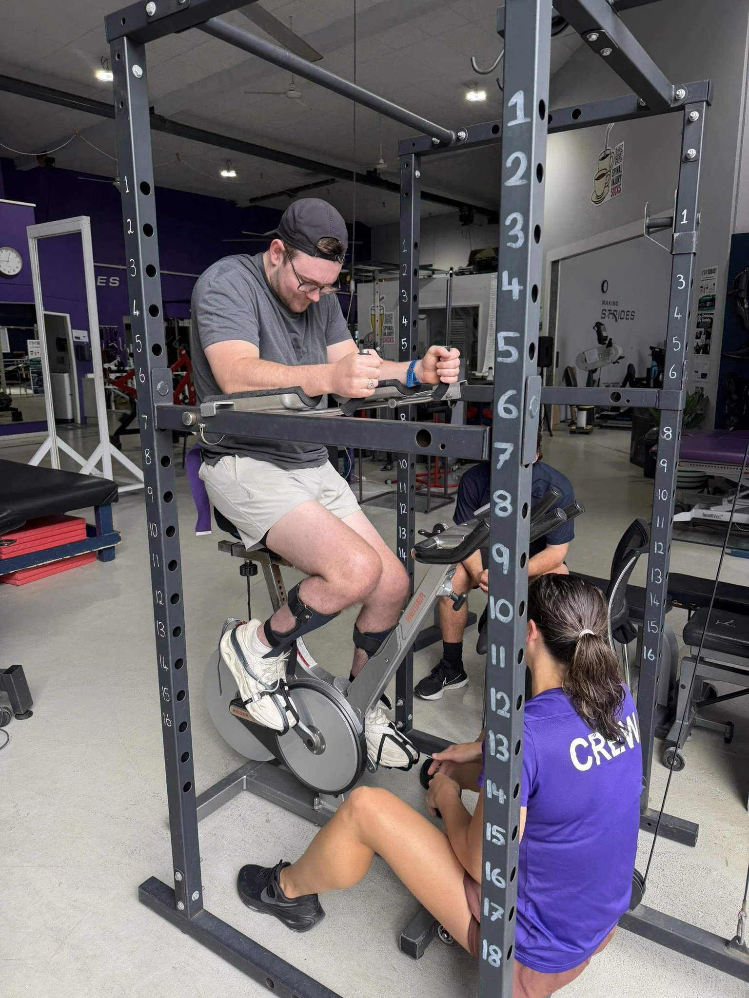 A woman in a purple shirt and a man in a gray shirt and black cap are working together on a stationary bike inside a gym. The man is adjusting the bike while the woman is kneeling beside him, assisting or observing.