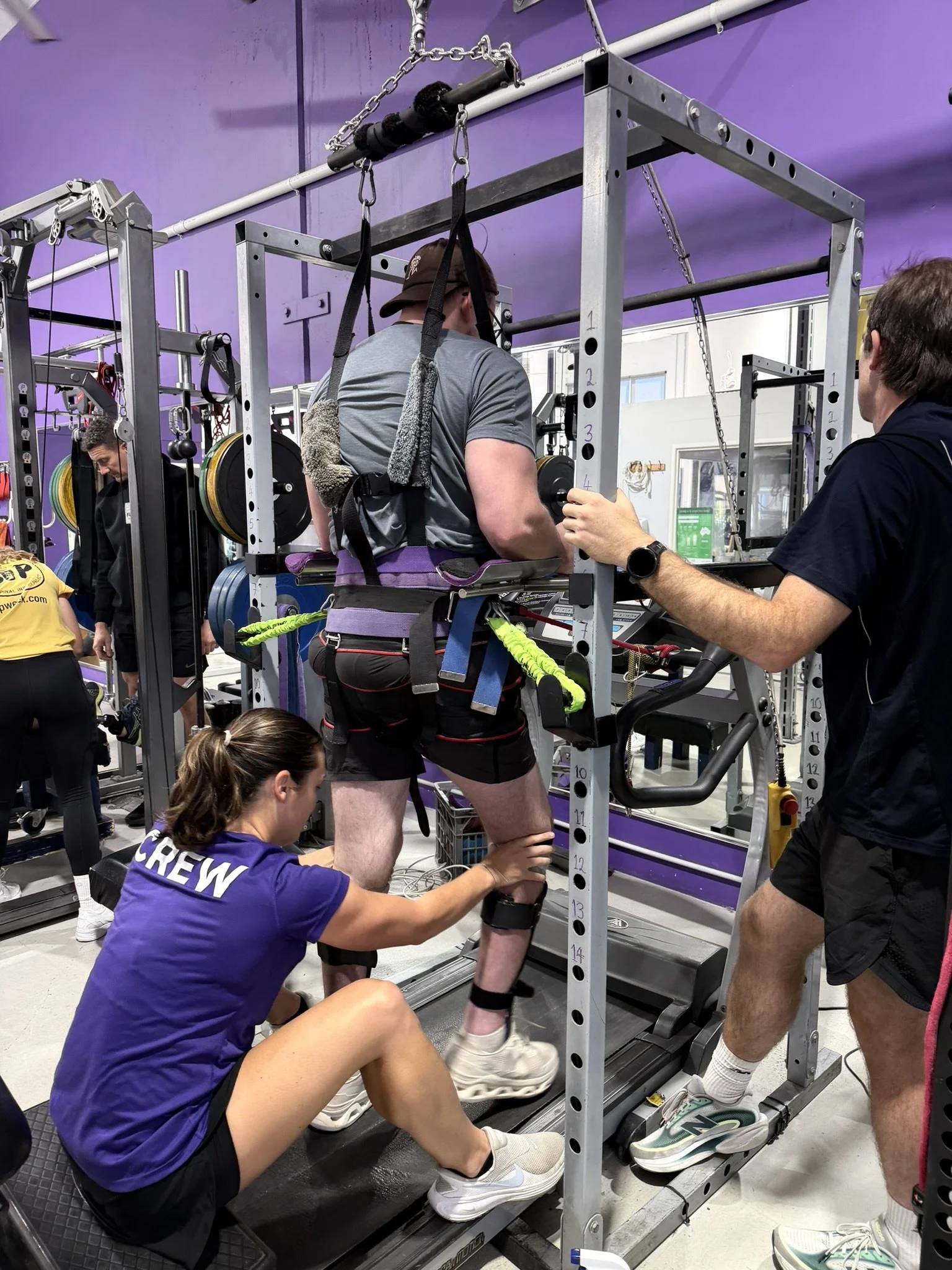 A person with a harness around their waist and a harness on their legs is walking on a treadmill with assistance from a woman with a purple shirt kneeling on the floor. The person is holding onto a bar with straps hanging above, and another person is standing nearby supervising. The setting is an indoor gym with purple walls.
