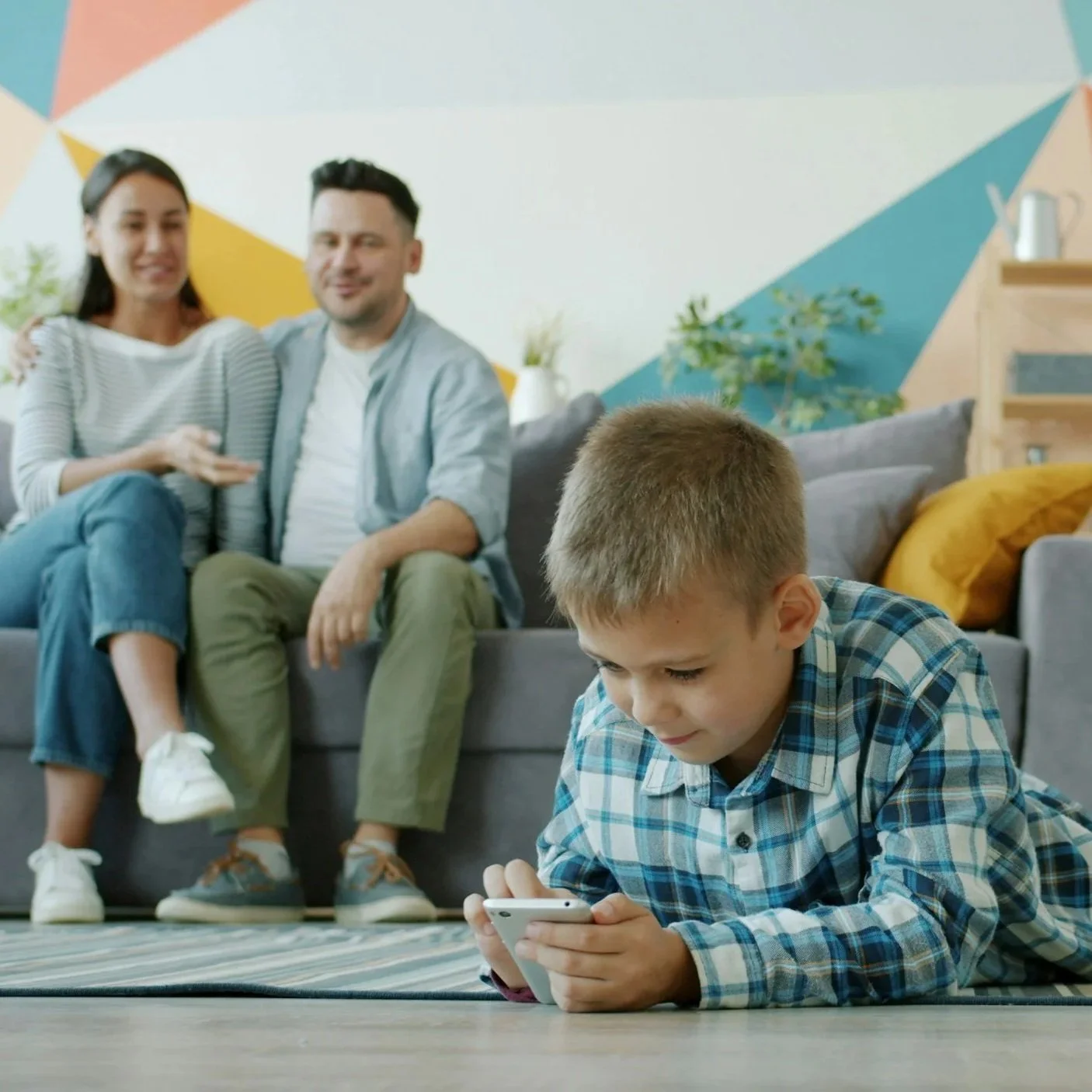 A boy lying on the floor playing on a smartphone, with a man and woman sitting on the couch behind him, smiling, in a brightly decorated living room.