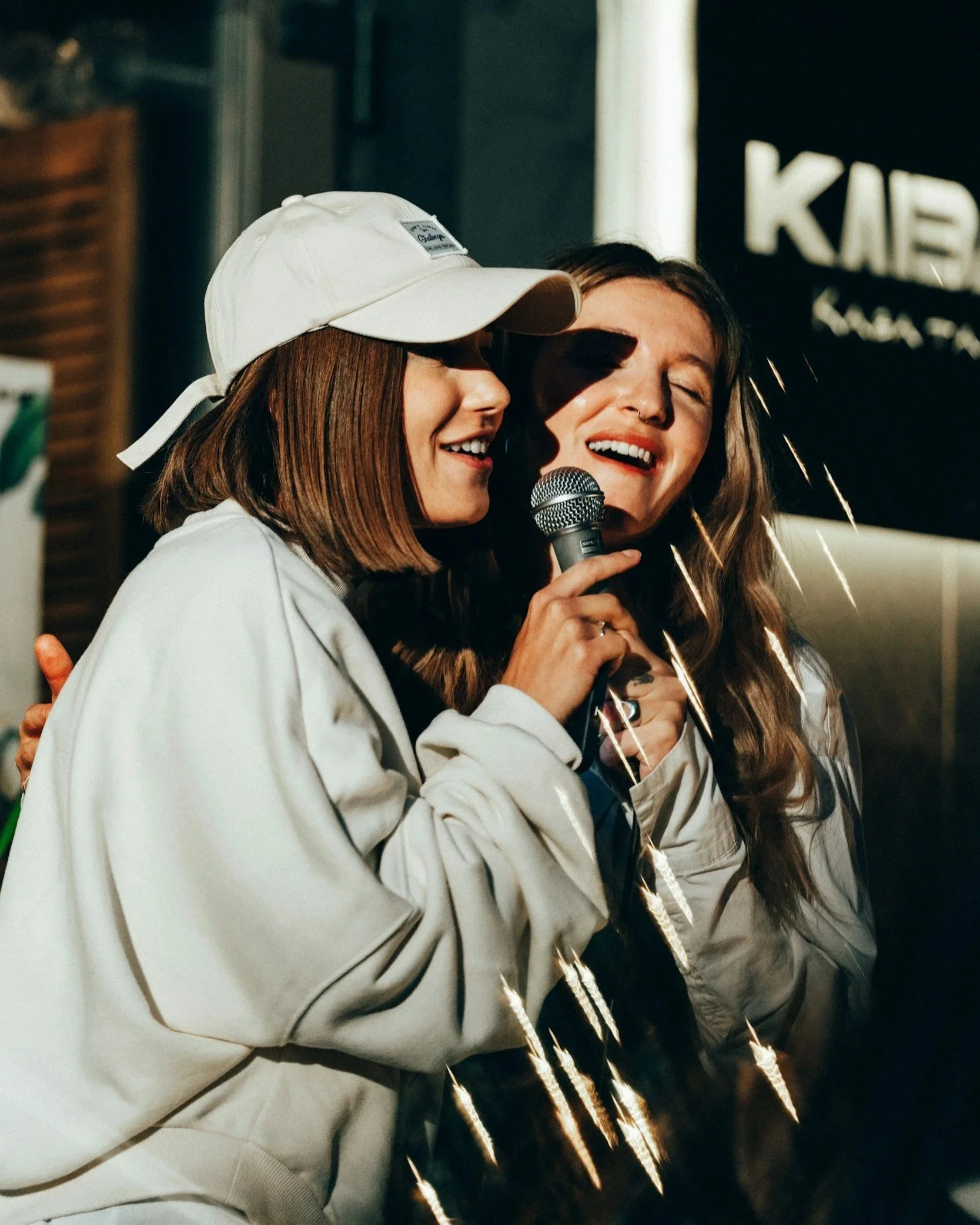 Two women laughing together, one holding a microphone, in a casual setting with sign and decorations in the background.