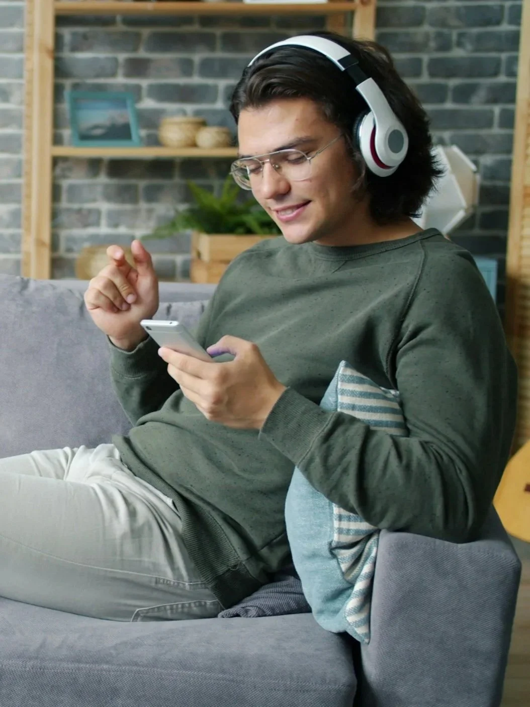 Young man sitting on a gray couch with a blue pillow, wearing headphones and glasses, looking at a smartphone while smiling.