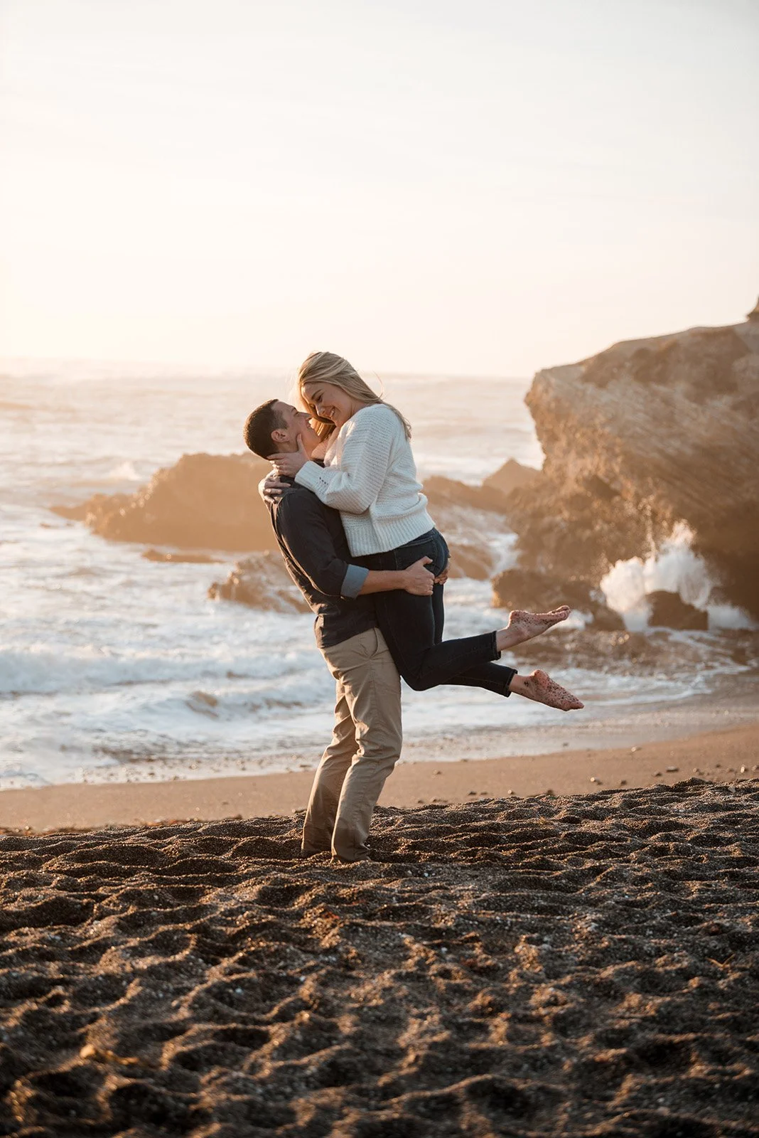 Lyndsi Seth Photography-A man lifting a woman in his arms on a sandy beach as waves crash against rocky cliffs in the background