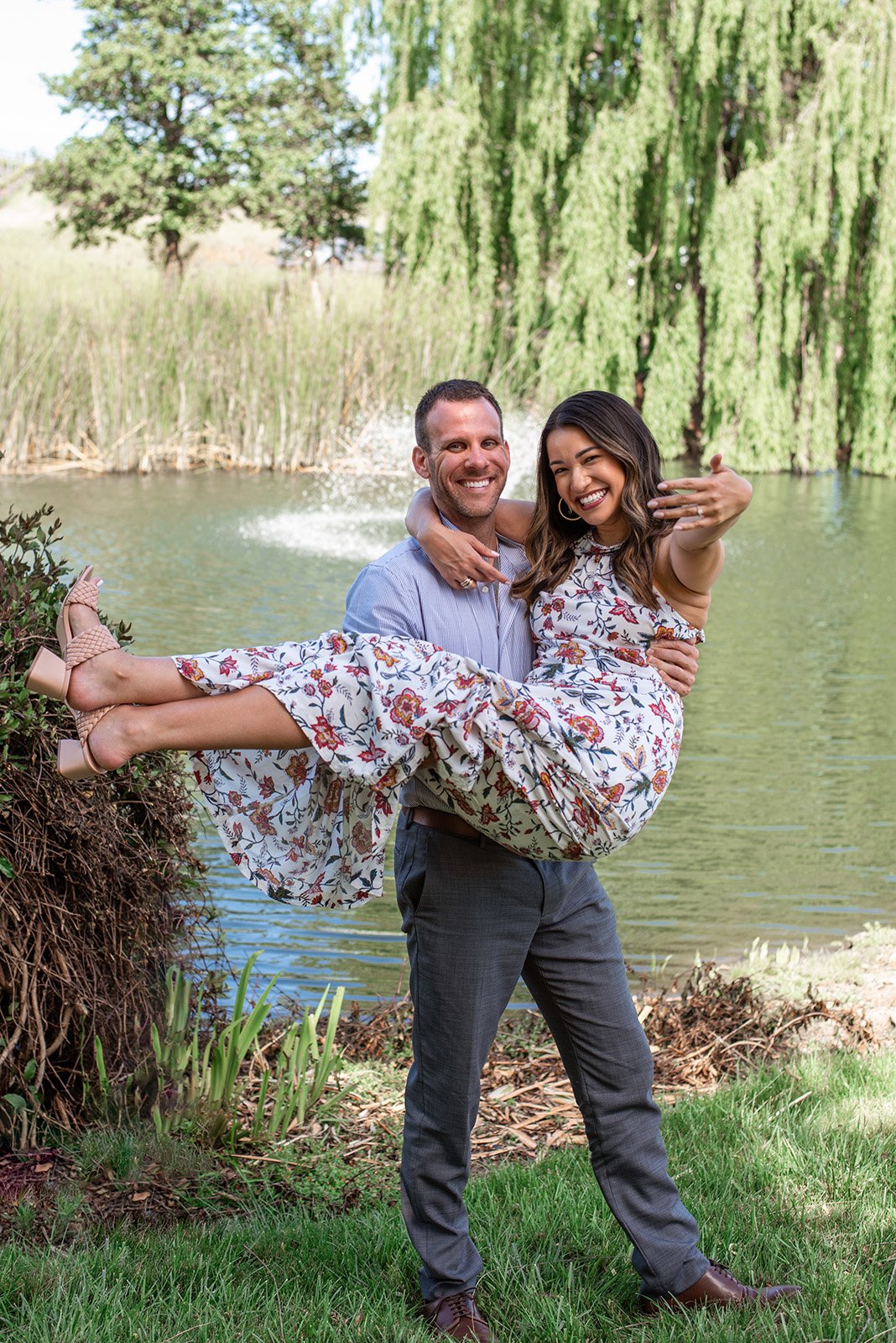 Lyndsi Seth Photography-A man carrying his partner outdoors in front of a pond as she excitedly shows her engagement ring.