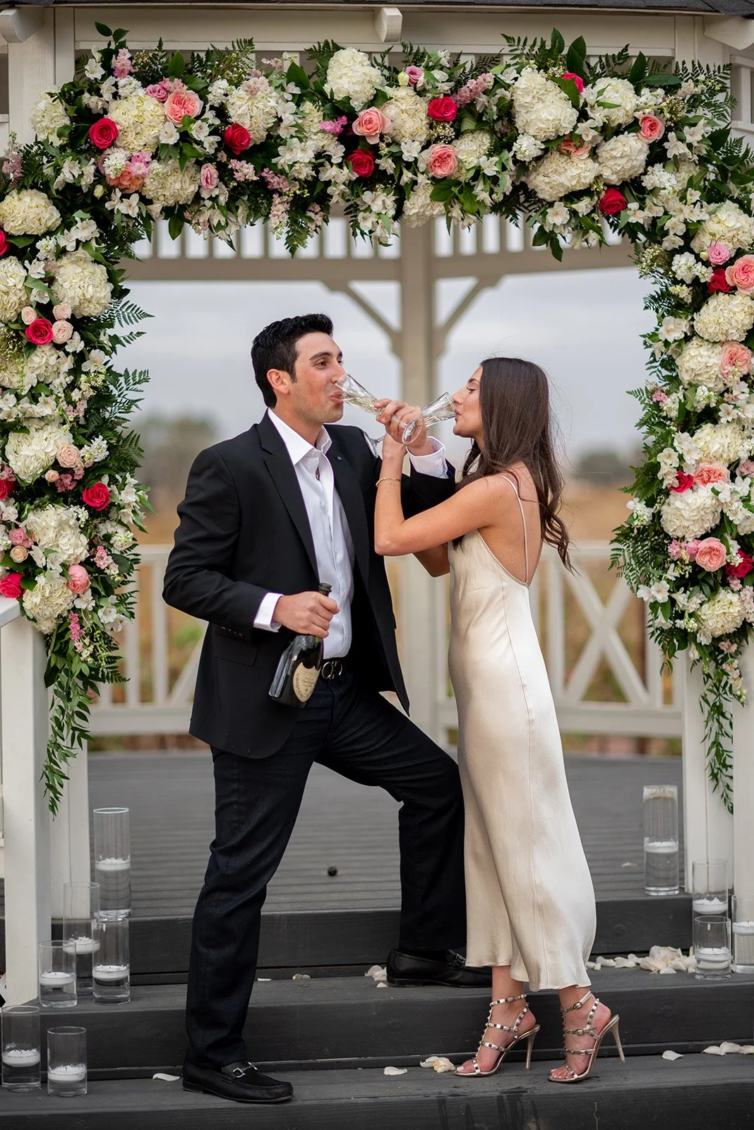 A couple at a wedding decorated with a floral arch, celebrating with drinks, standing on a black deck with water glasses, and rose petals scattered on the ground.