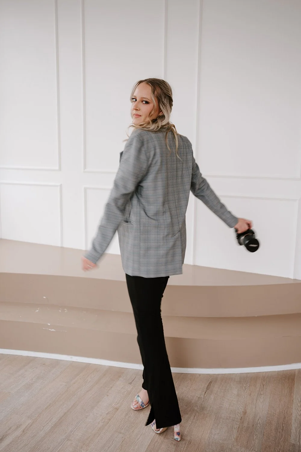 A woman in a gray plaid blazer and black pants holding a camera in a white room.