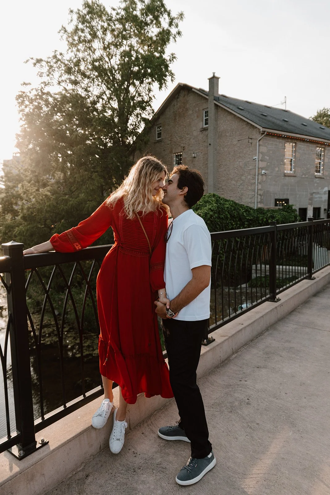 Lyndsi Seth Photography-A smiling couple holding hands outdoors during an engagement photoshoot, with the woman wearing a long red dress and white sneakers, and the man wearing a white shirt and dark pants
