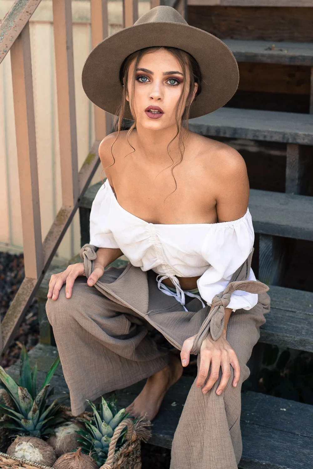 A young woman wearing a wide-brimmed hat, off-shoulder white blouse, and loose khaki pants, squatting on outdoor steps near a basket of pineapple plants.
