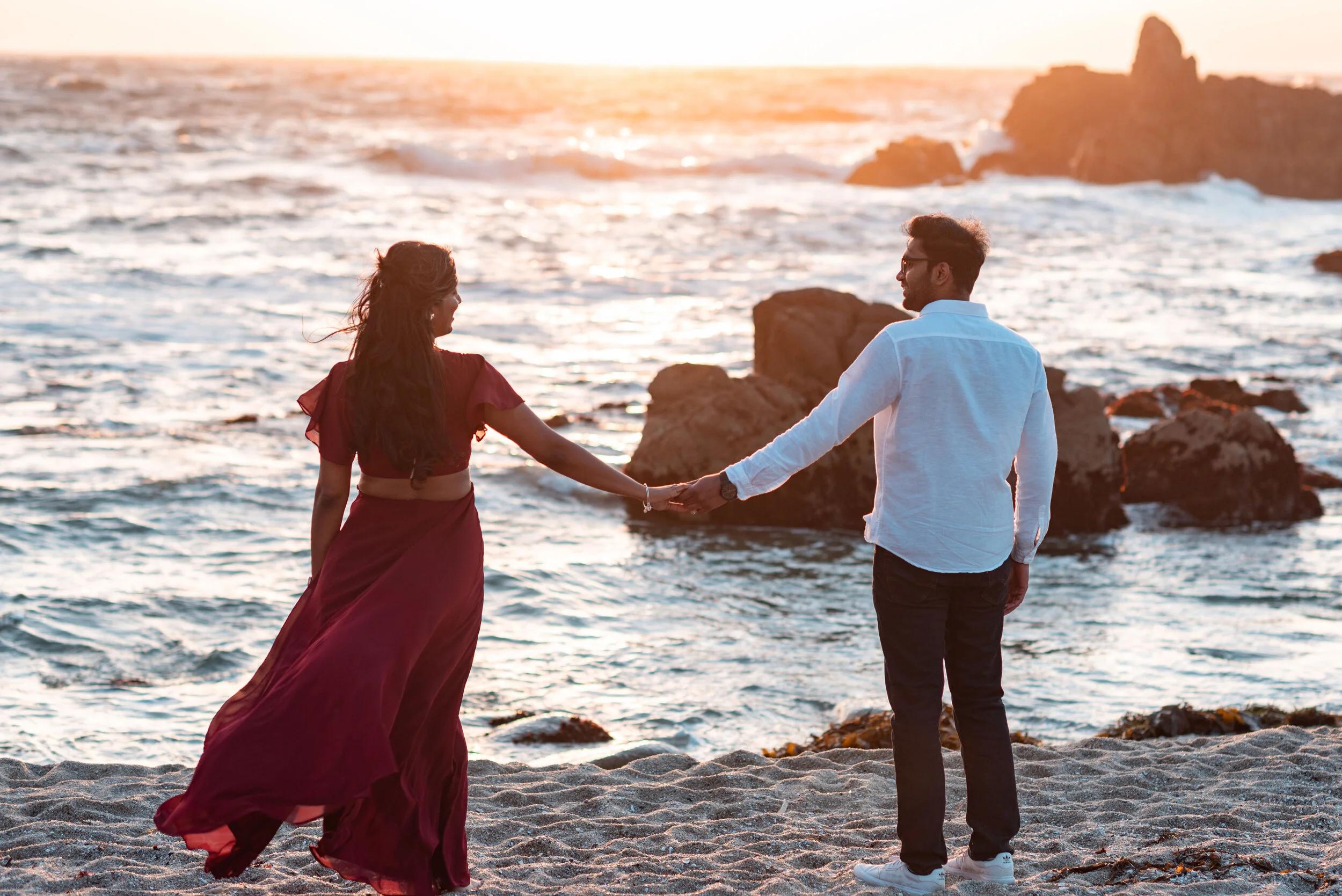 A man and woman holding hands facing each other on a beach at sunset, with ocean waves and rocks in the background.