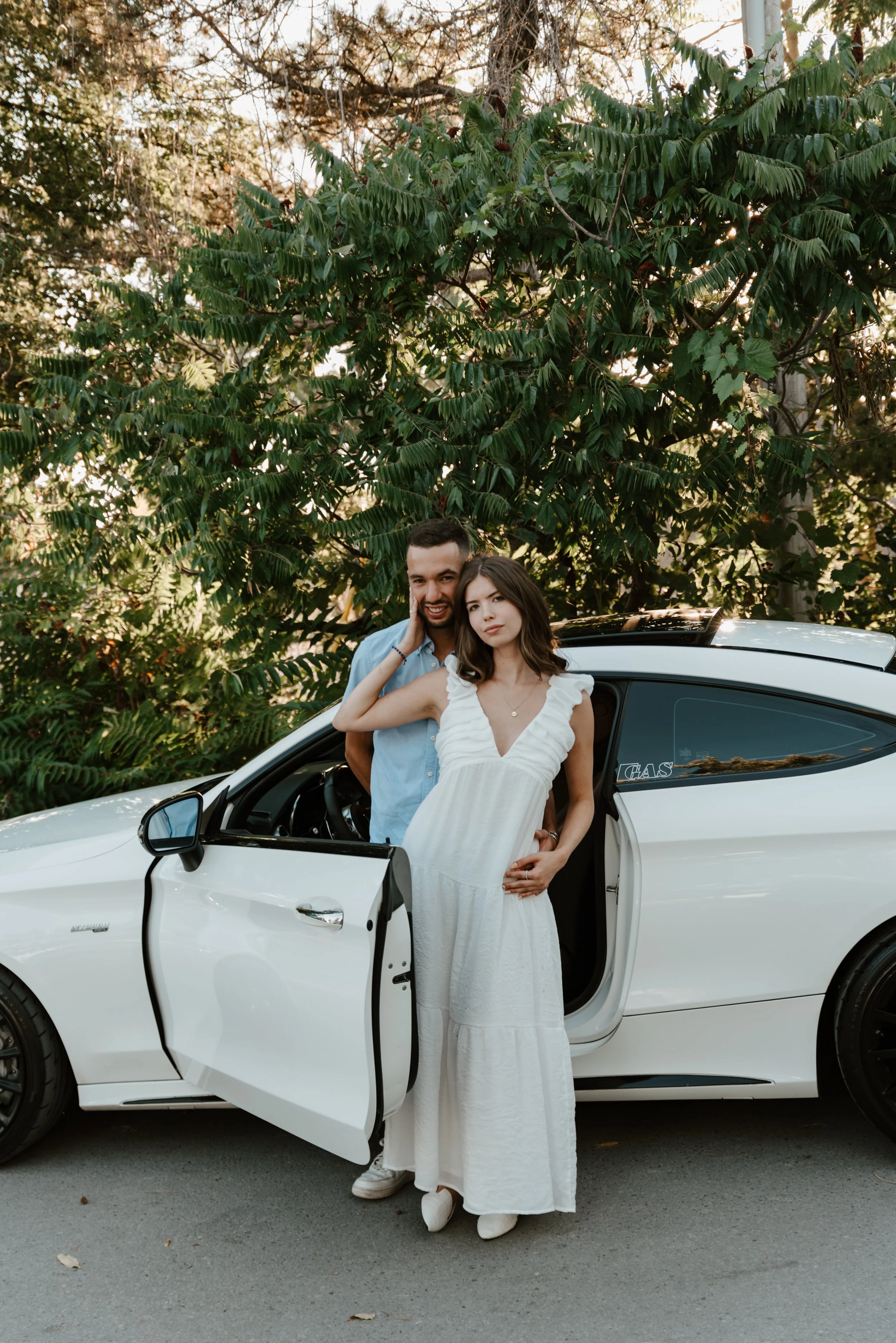 A young couple stands in front of a white electric car with its door open, surrounded by lush green trees. The man, dressed in a light blue shirt, and the woman, in a white dress, pose for a photo during daylight.