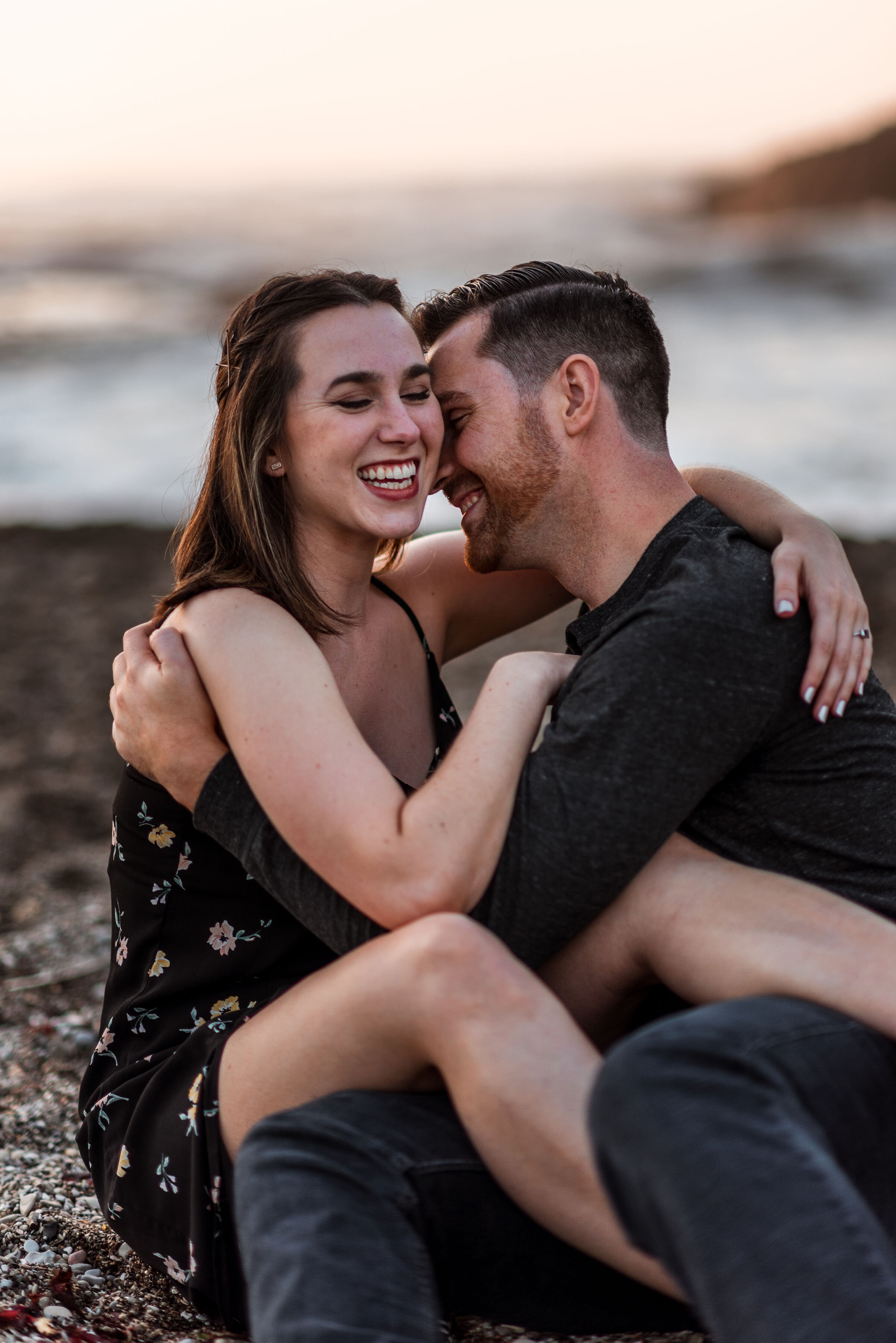 A smiling couple embracing each other on a beach at sunset, with the woman wearing a floral dress and the man wearing a dark shirt.
