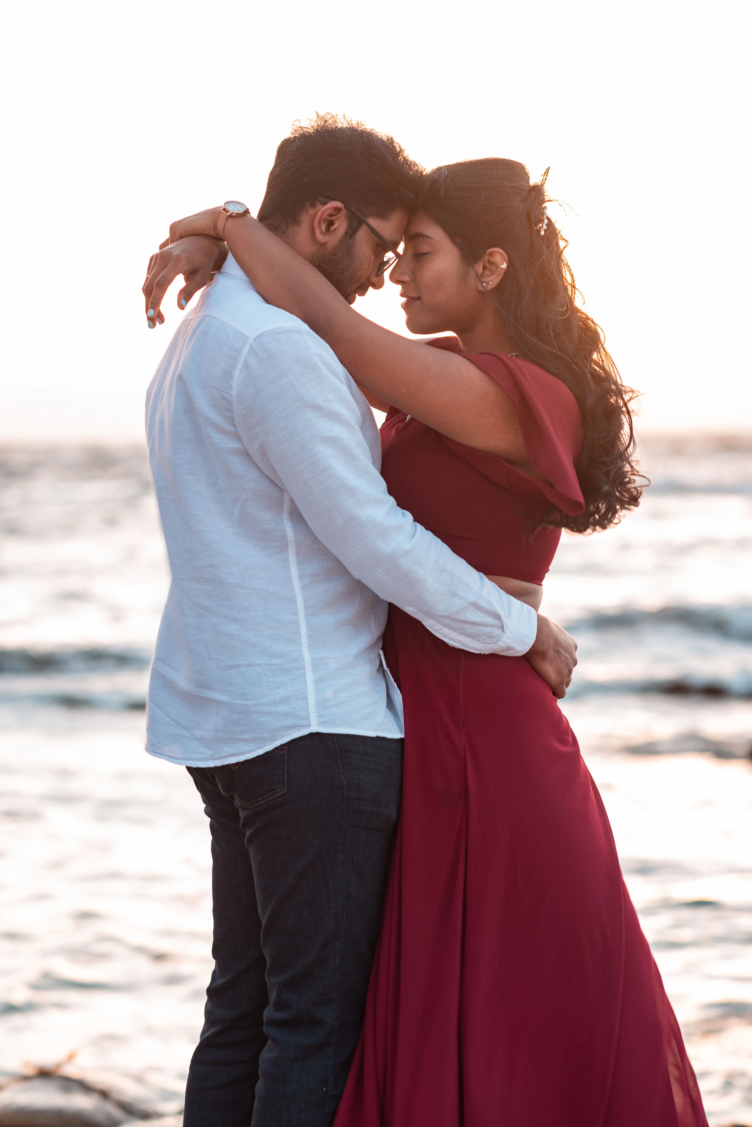 A couple embracing on the beach at sunset with their foreheads touching, eyes closed, and calm expressions, in both casual and romantic mood.