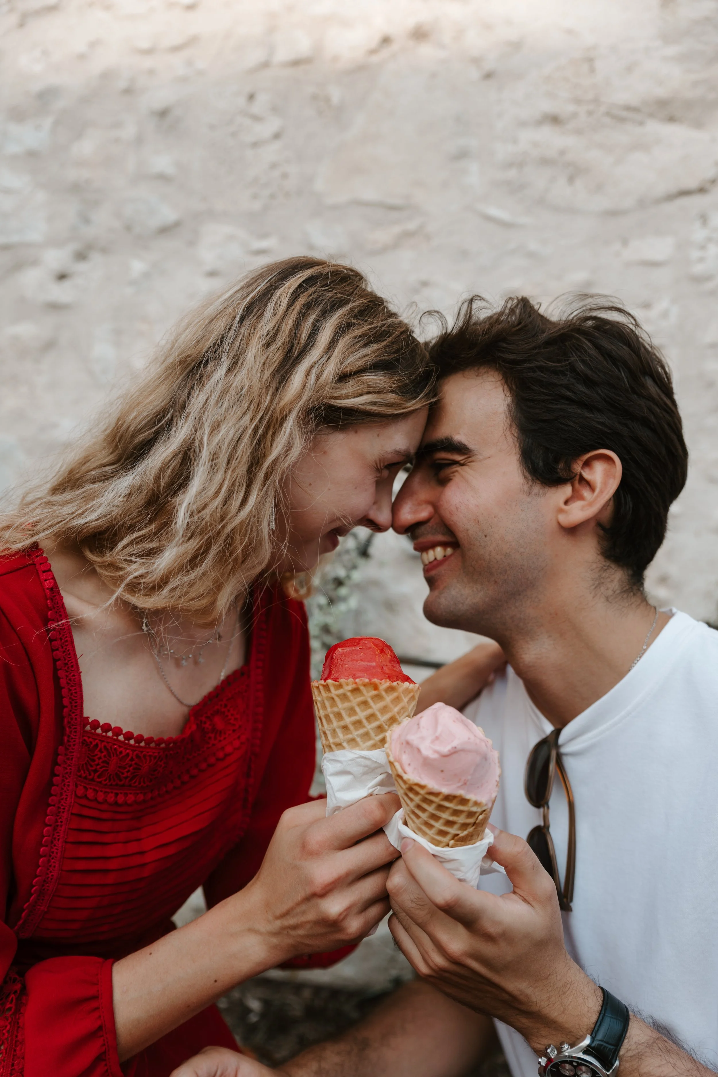 A couple sharing a moment with their foreheads touching while holding two ice cream cones, smiling, against a stone wall background.