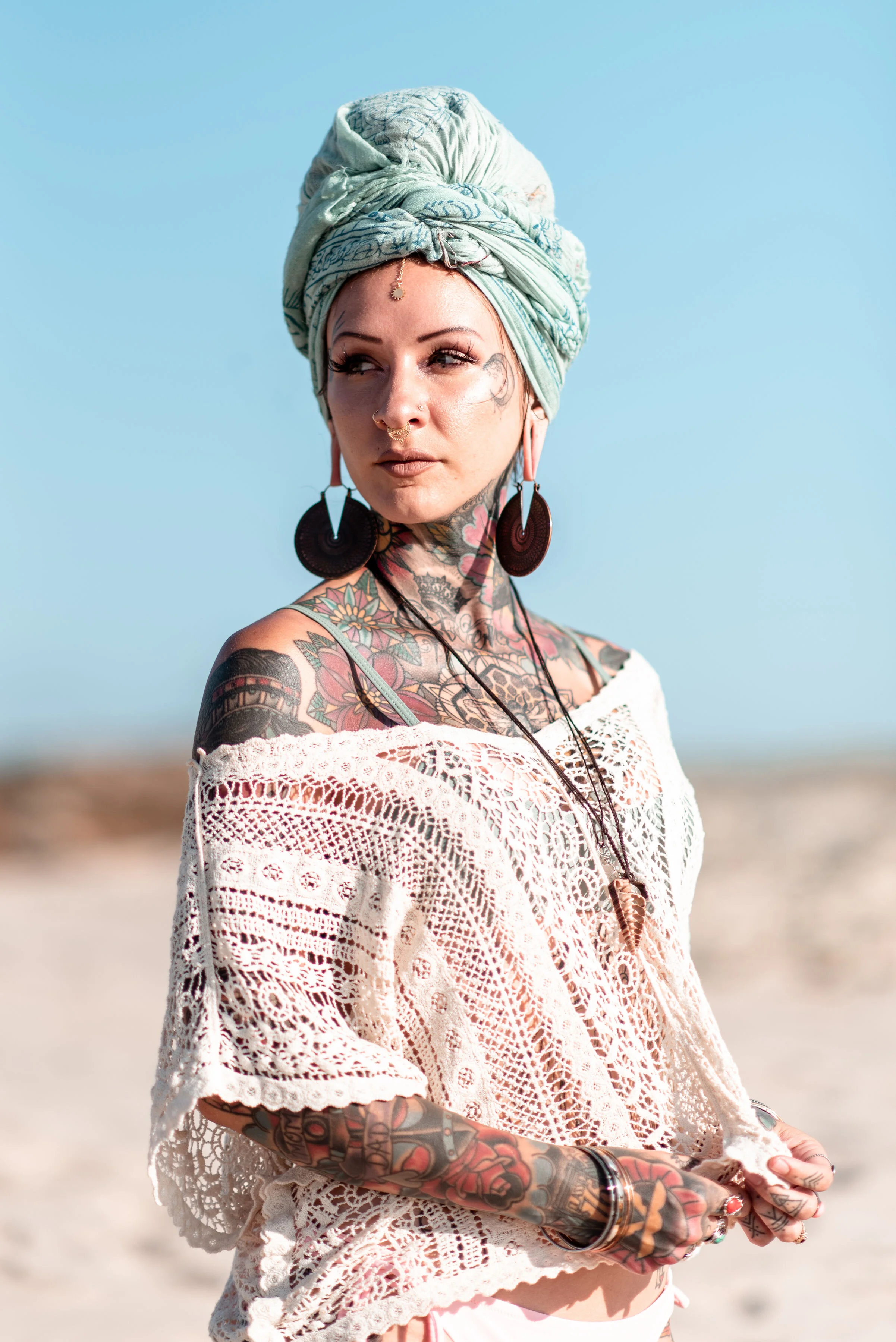 A woman on the beach with tattoos, wearing a light-colored crochet top, large earrings, and a headwrap, looking to the side with a thoughtful expression.