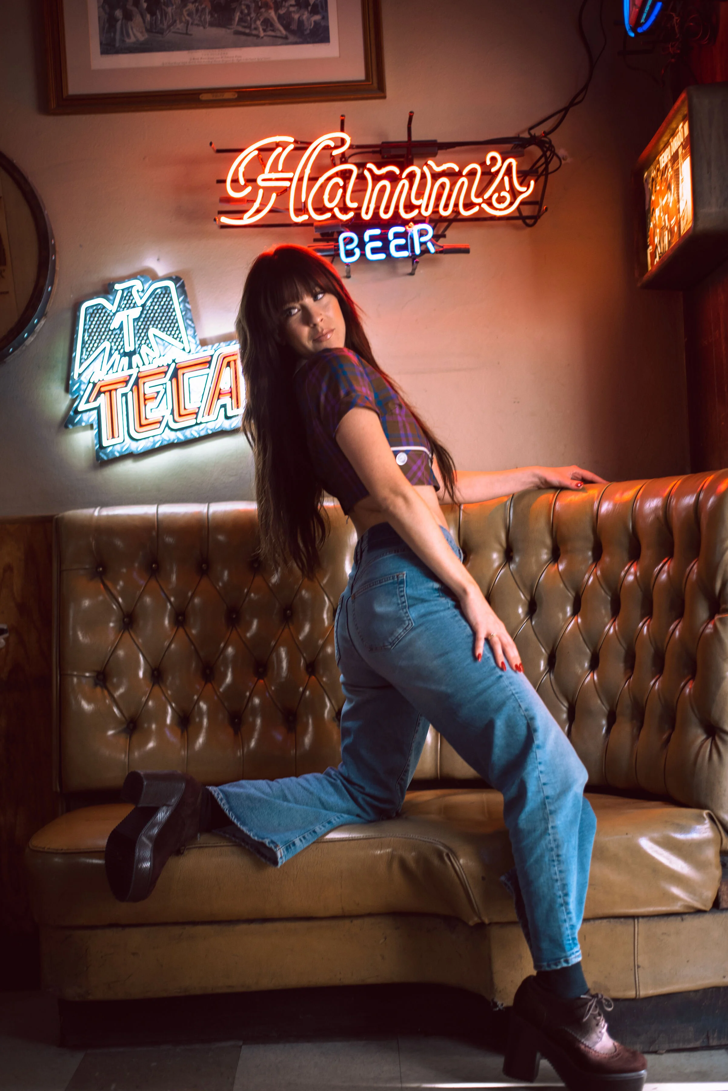 A woman with long dark hair in plaid clothing and jeans poses on a leather booth in a bar or restaurant. She is kneeling with her left knee on the seat, her right foot on the floor, and her right hand resting on the back of the booth. Behind her, there are neon signs that read 'Hamms Beer' and 'Texas', along with a framed picture on the wall.