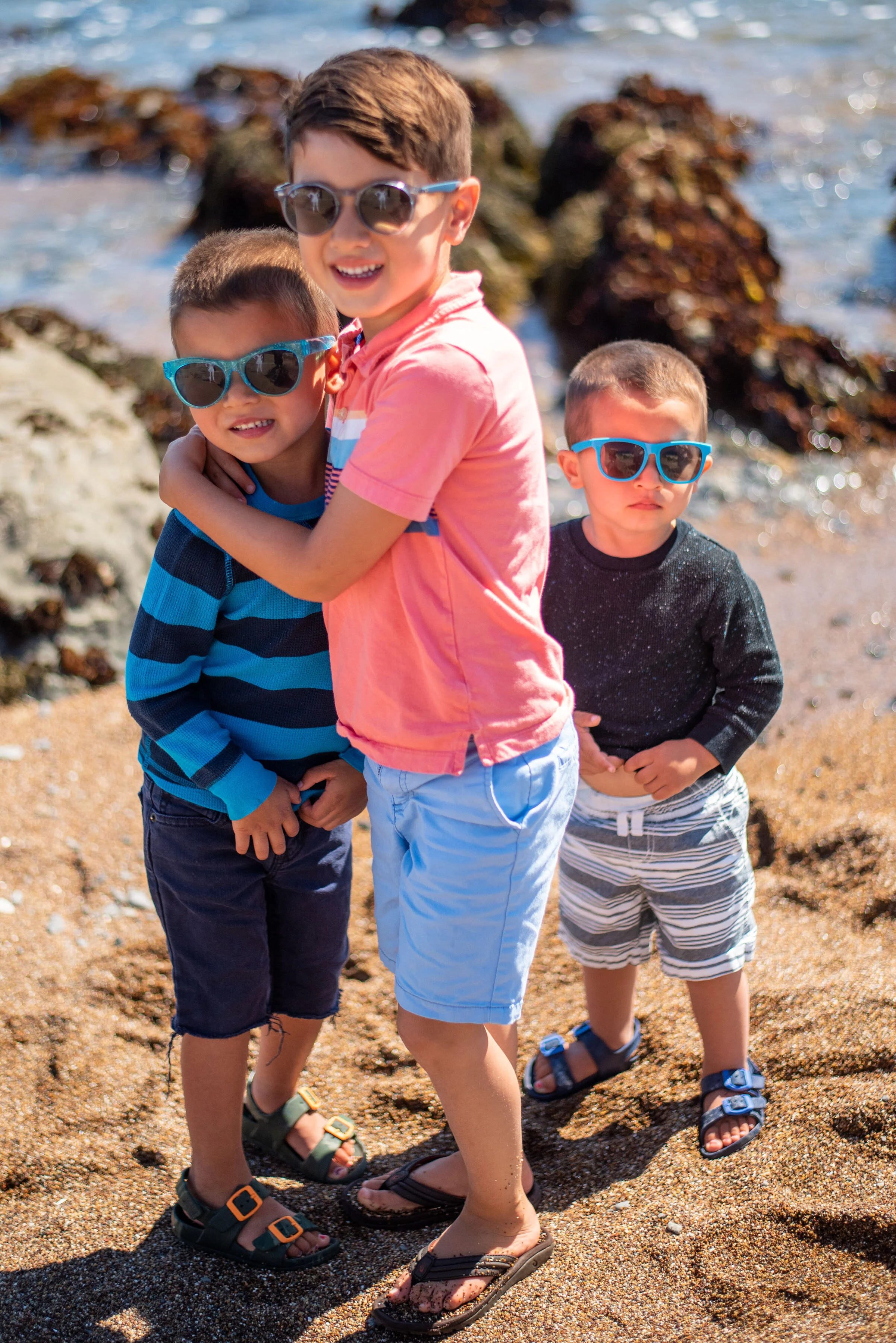 Four young boys wearing sunglasses standing on a sandy beach near the water with rocks in the background. They are smiling and dressed in casual summer clothes with sandals.