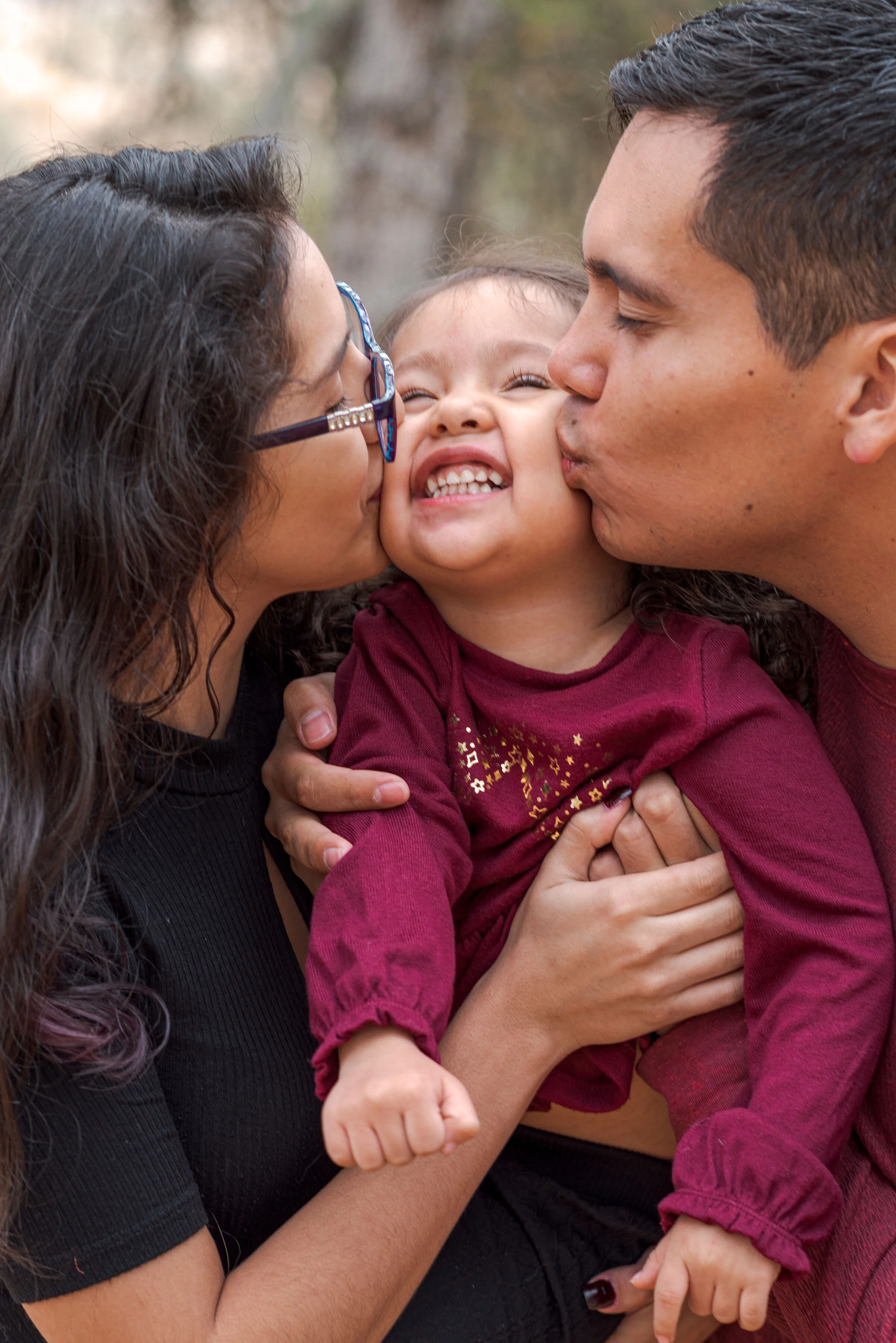 A family of three, parents and a child, embracing and sharing kisses outdoors during daytime.