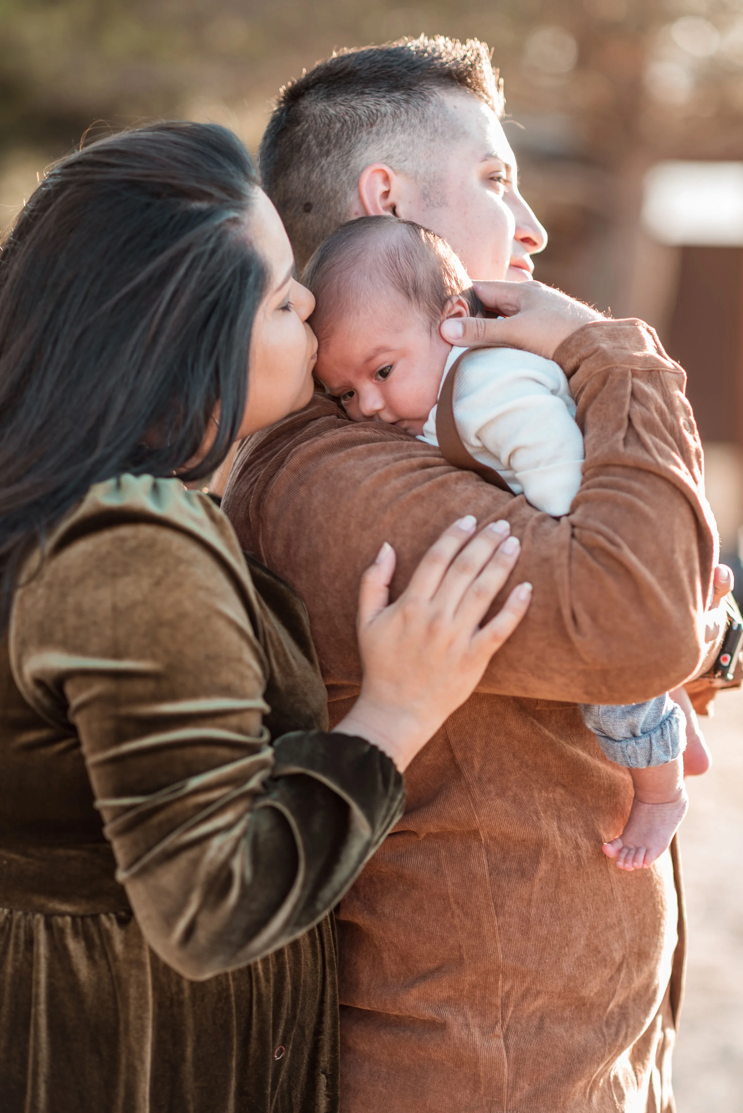A woman is kissing a newborn baby on the forehead while a man holds the baby, all outdoors with sunlight in the background.