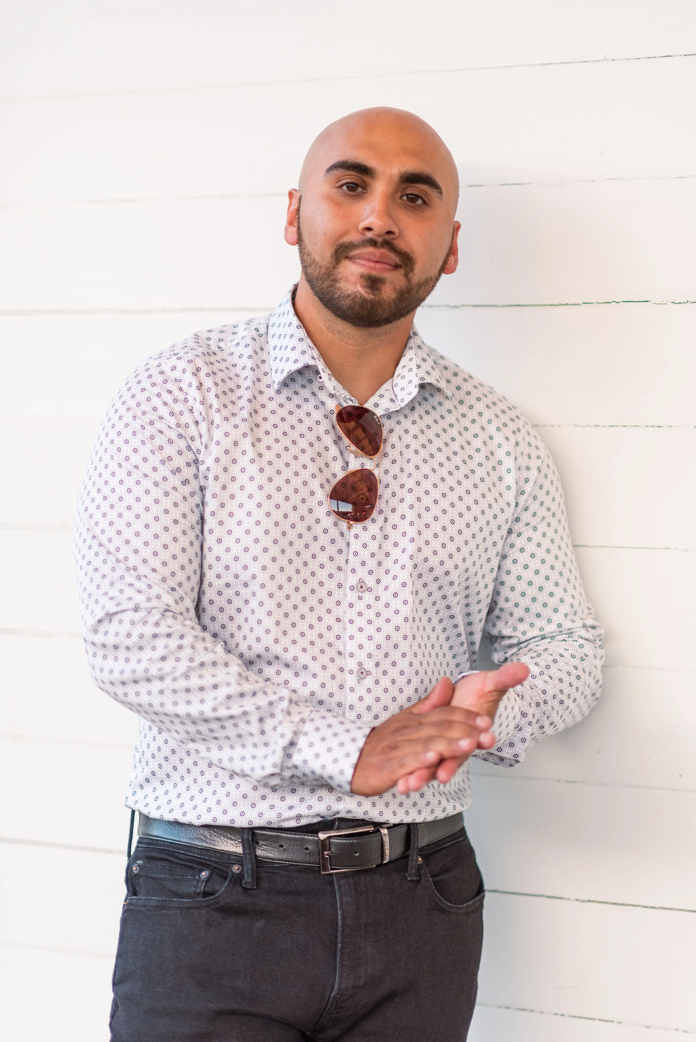 A man with a shaved head and beard wearing a white patterned shirt with sunglasses hanging from the collar, standing against a white wooden wall.