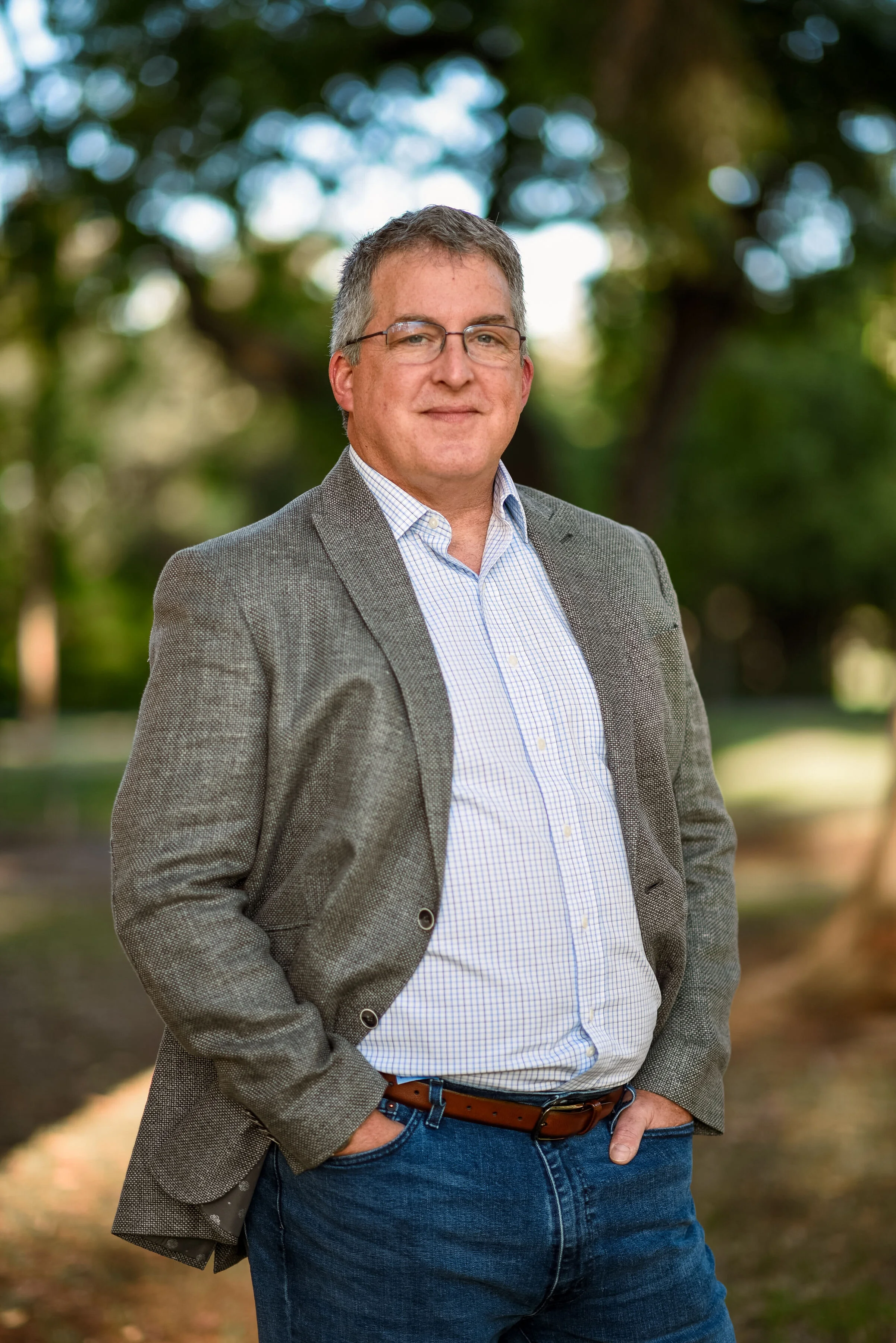 A man with glasses and short, gray hair stands outdoors with trees blurred in the background. He is wearing a gray blazer over a light checkered shirt with jeans, and is smiling slightly with one hand in his pocket.