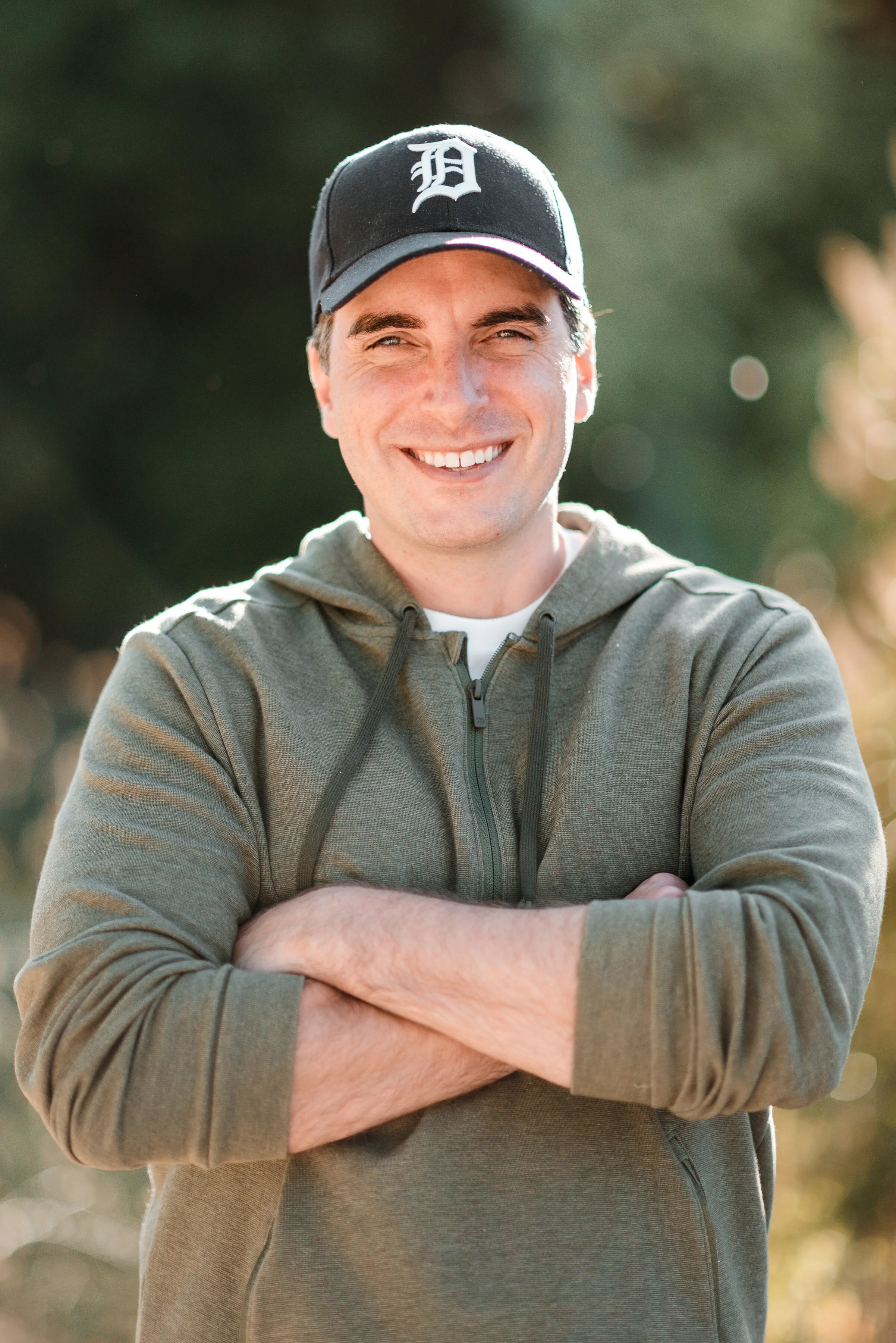 A young man with light skin, short brown hair, and a big smile, wearing a black baseball cap with a white logo, a gray zip-up hoodie, and standing outdoors with blurred trees in the background.