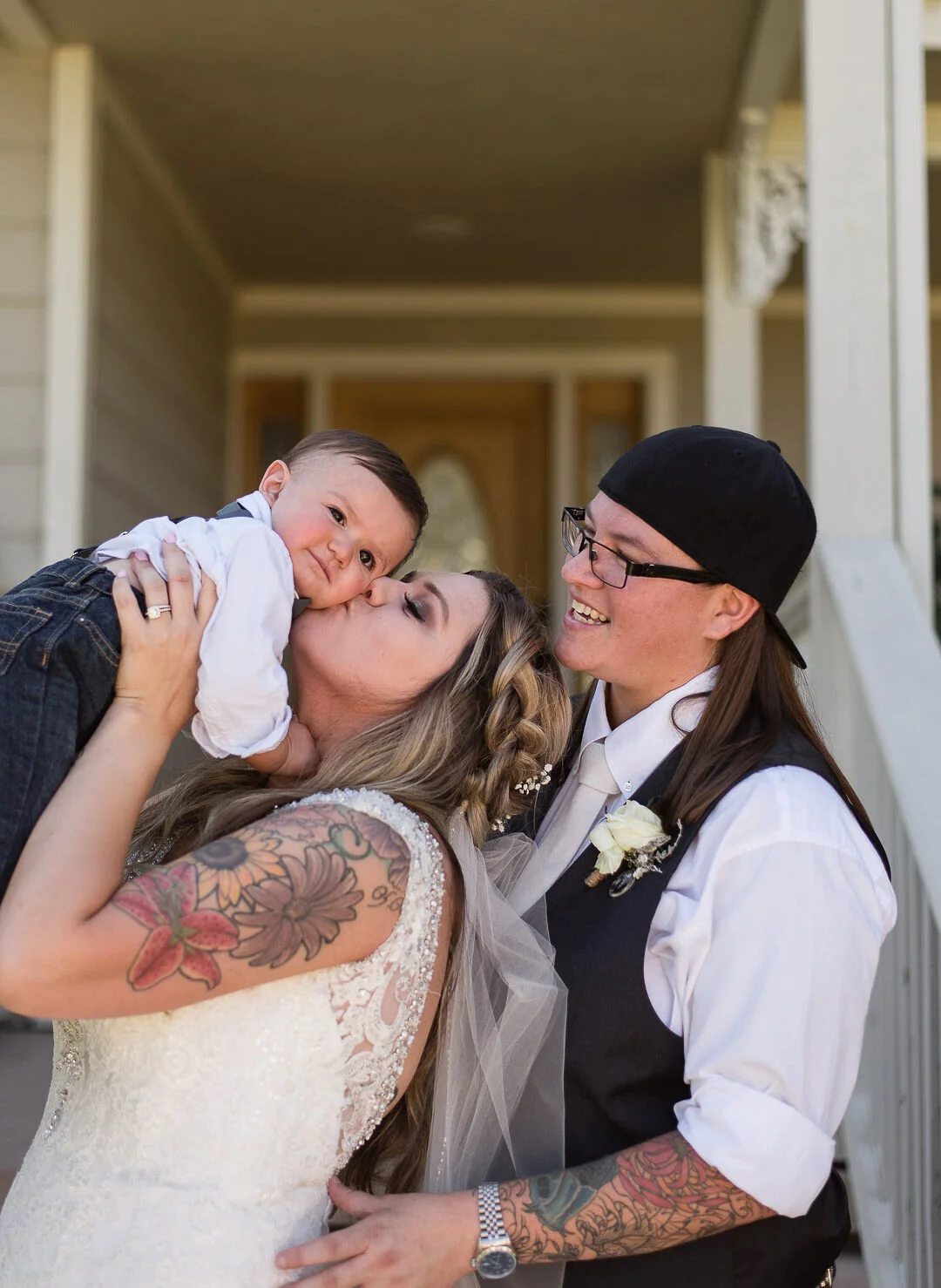 A newlywed couple with tattoos, holding a young boy, who is leaning in to give the bride a kiss on the cheek, outdoors in front of a house.