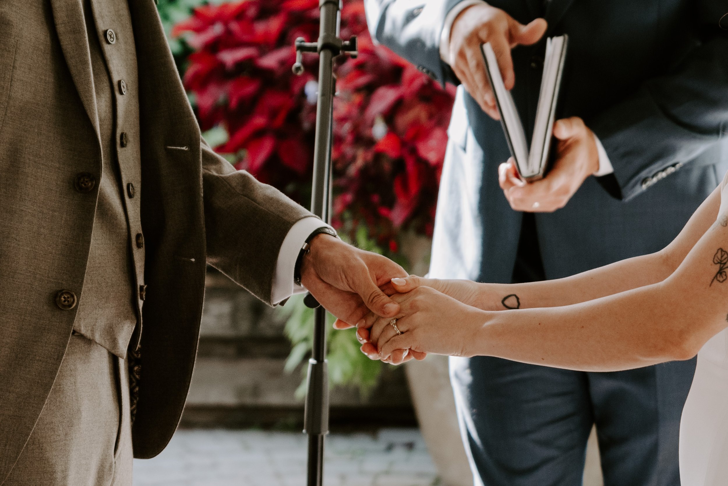 A couple holding hands during a wedding ceremony, with the bride's tattooed arm visible, and someone in the background holding a folder or book.