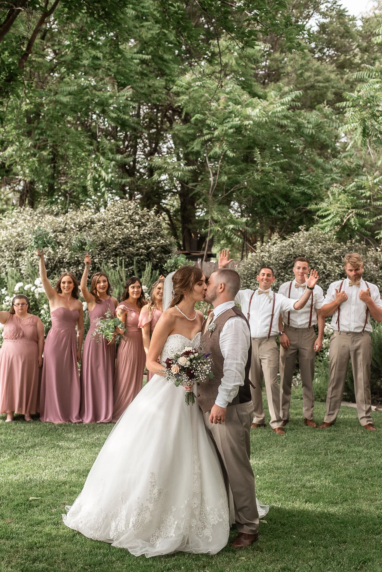 A bride and groom kissing outdoors, surrounded by bridesmaids and groomsmen on a grassy area with trees in the background.