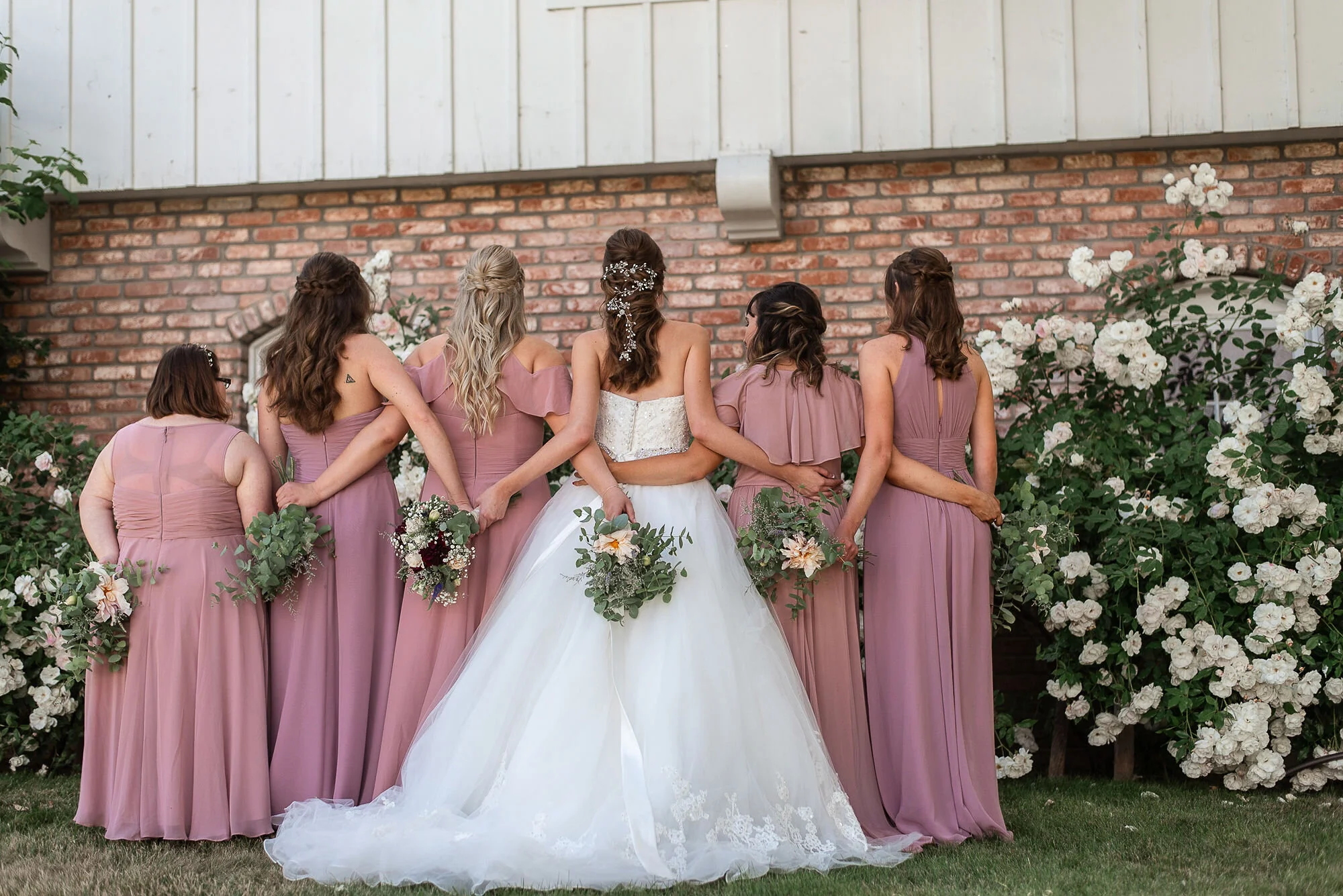 Lyndsi Seth photography- A bride in a ballgown standing back-to-camera with her bridesmaids in dusty rose dresses, holding bouquets behind their backs
