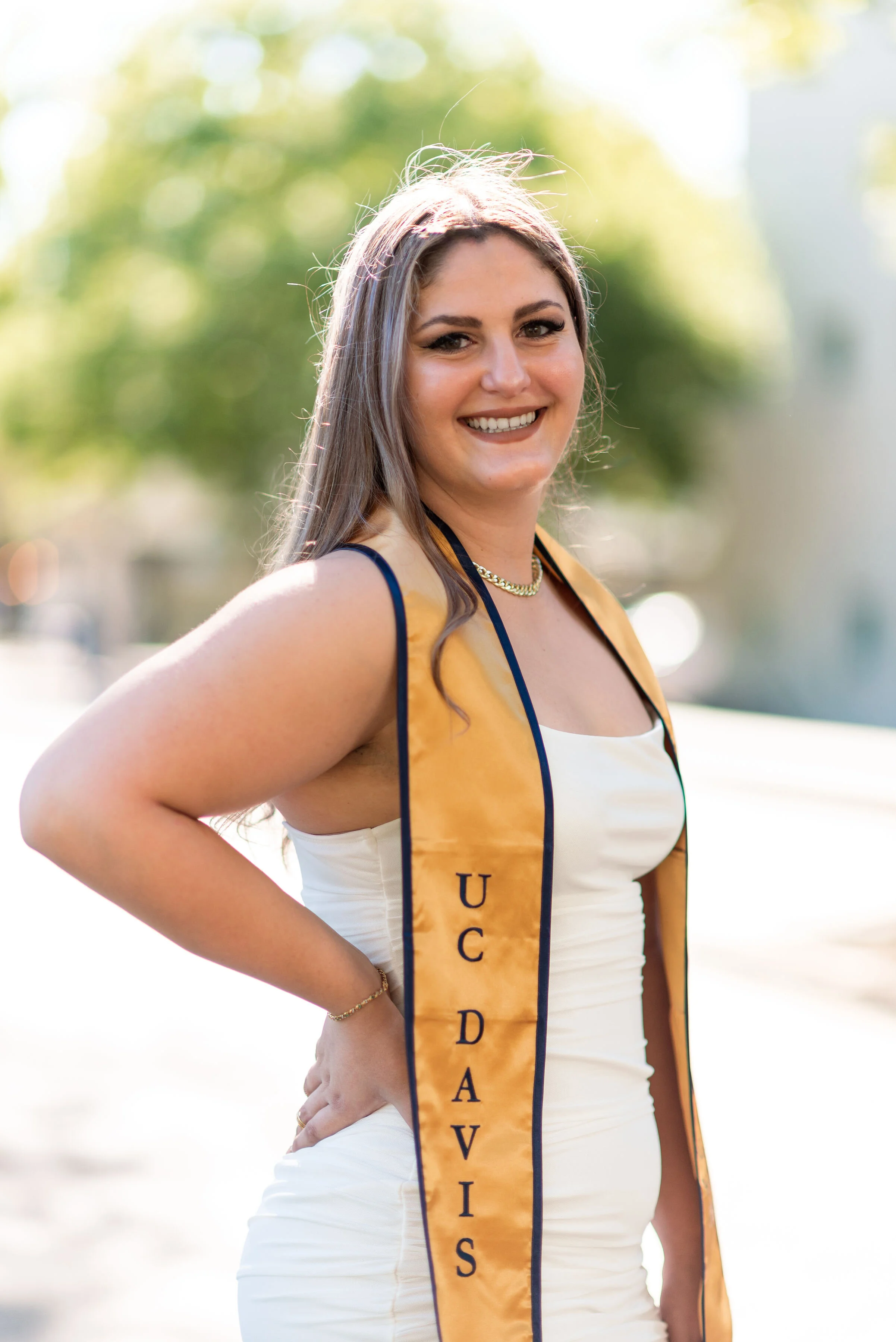 A young woman with long hair, smiling, wearing a cream dress and gold necklace, standing outdoors in sunlight with a yellow sash that has black text.