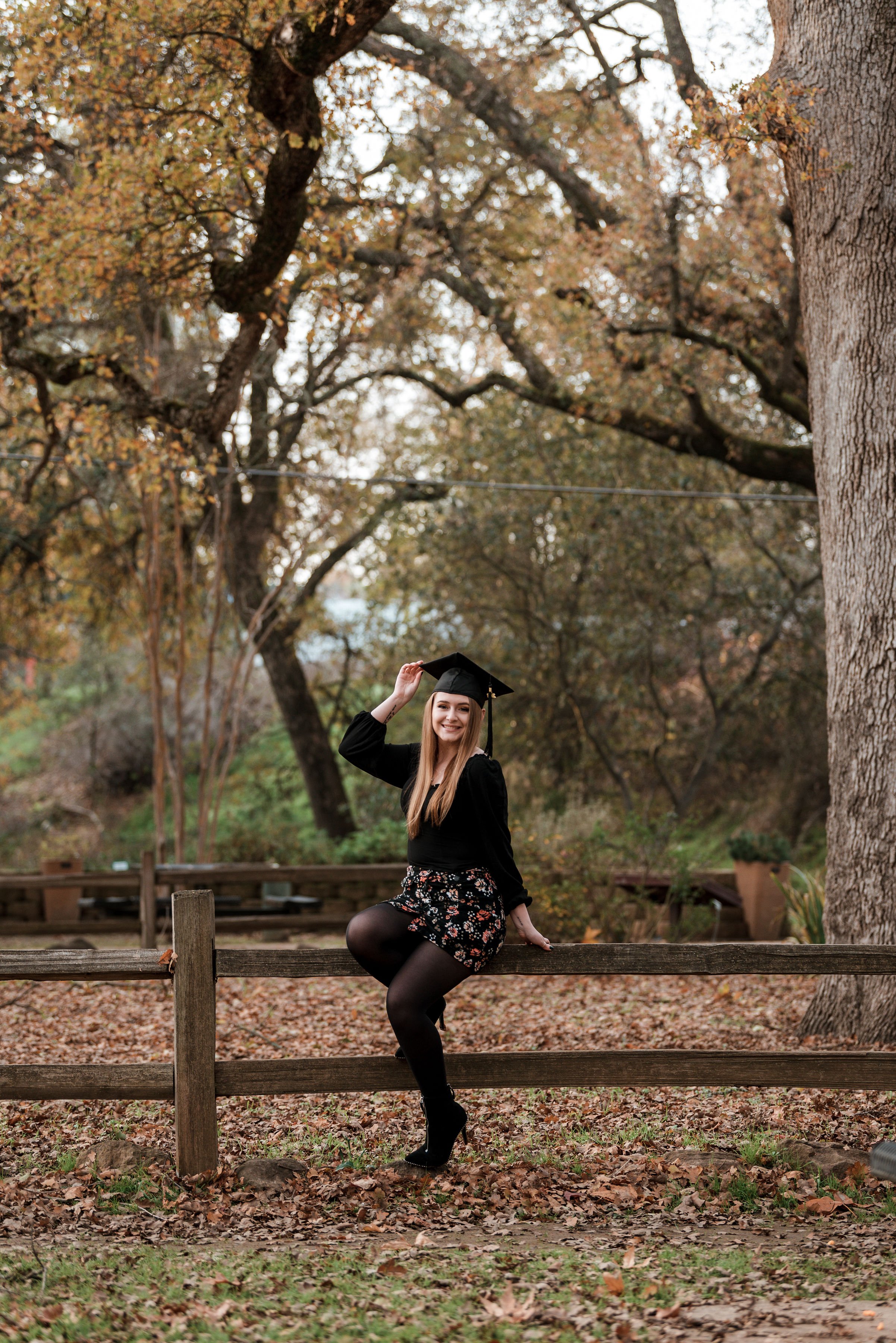 A young woman in a black floral skirt, black top, and black tights, sitting on a wooden fence in a park with autumn leaves. She is holding a graduation cap in her right hand and smiling at the camera. Large trees with autumn foliage surround her.