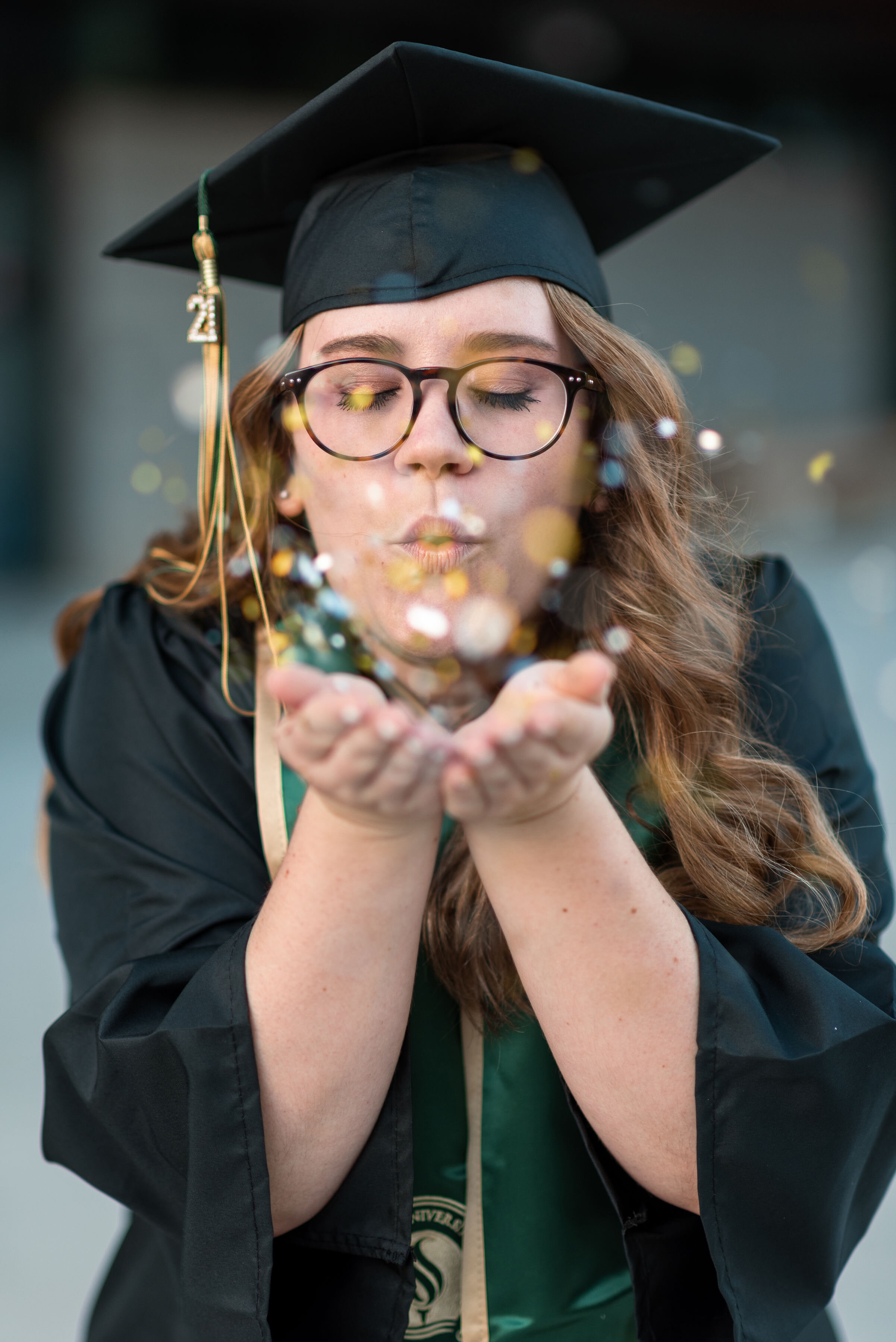 A young woman with glasses wearing a graduation cap and gown blows confetti from her hands with her eyes closed.