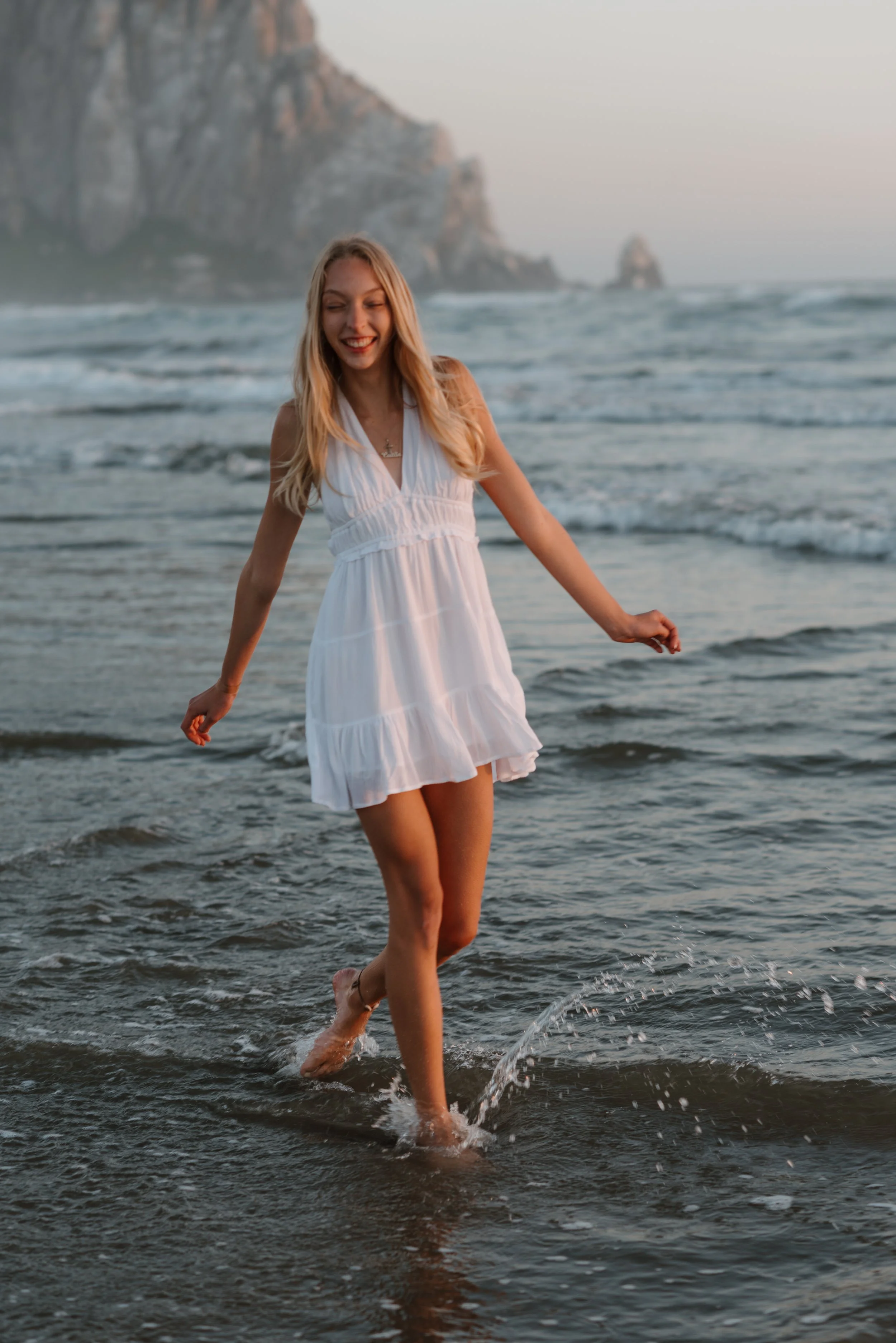 A woman in a white dress walking through shallow ocean water at sunset, with rocky cliffs in the background.