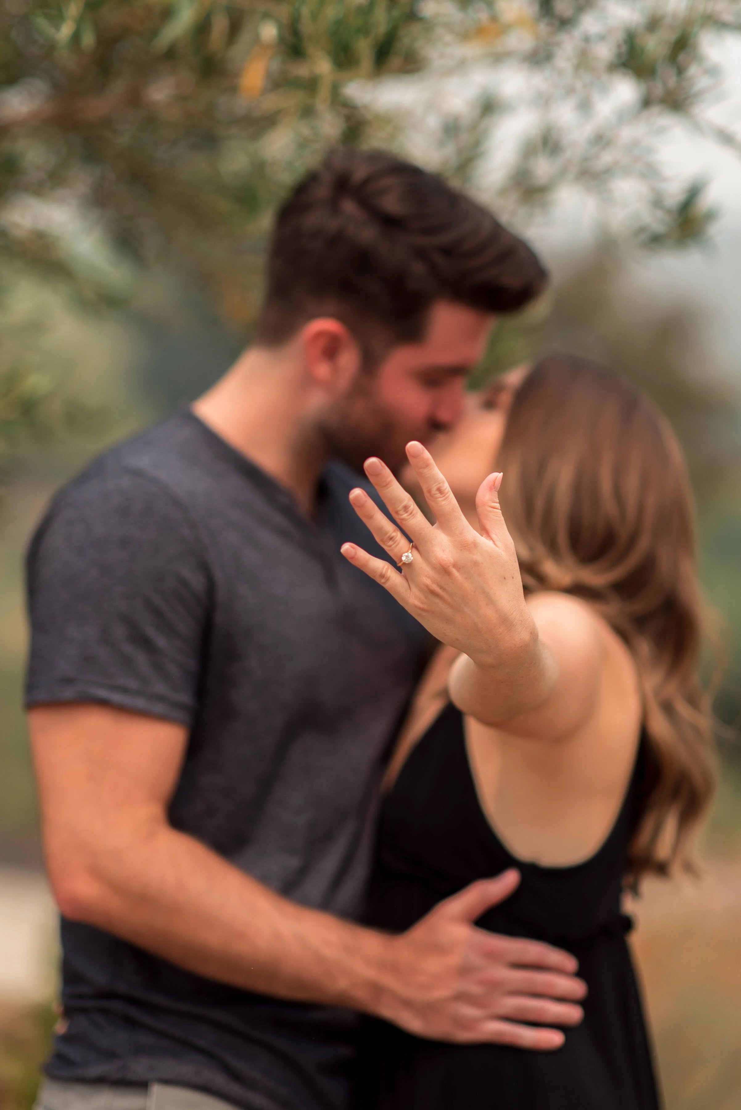 A close-up of a man and woman embracing outdoors, with the woman's left hand showing a diamond engagement ring, both with their faces close and blurred background of green foliage.