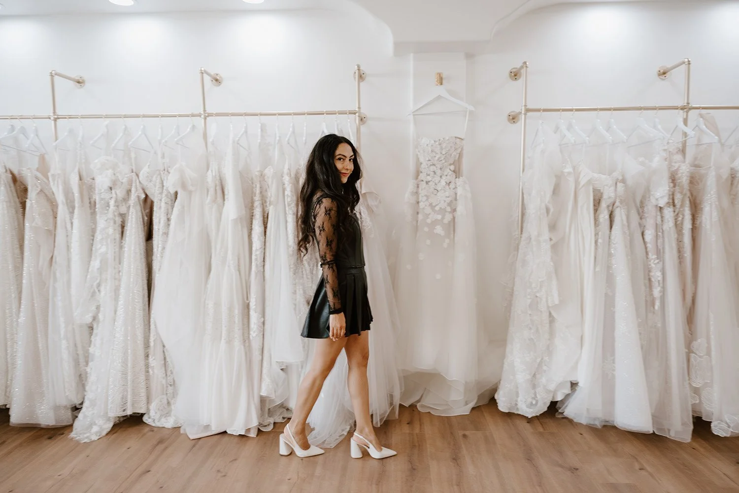 A woman with long dark hair in a black lace top and leather skirt shopping for wedding dresses in a bridal shop, standing in front of a display of white wedding gowns on racks.